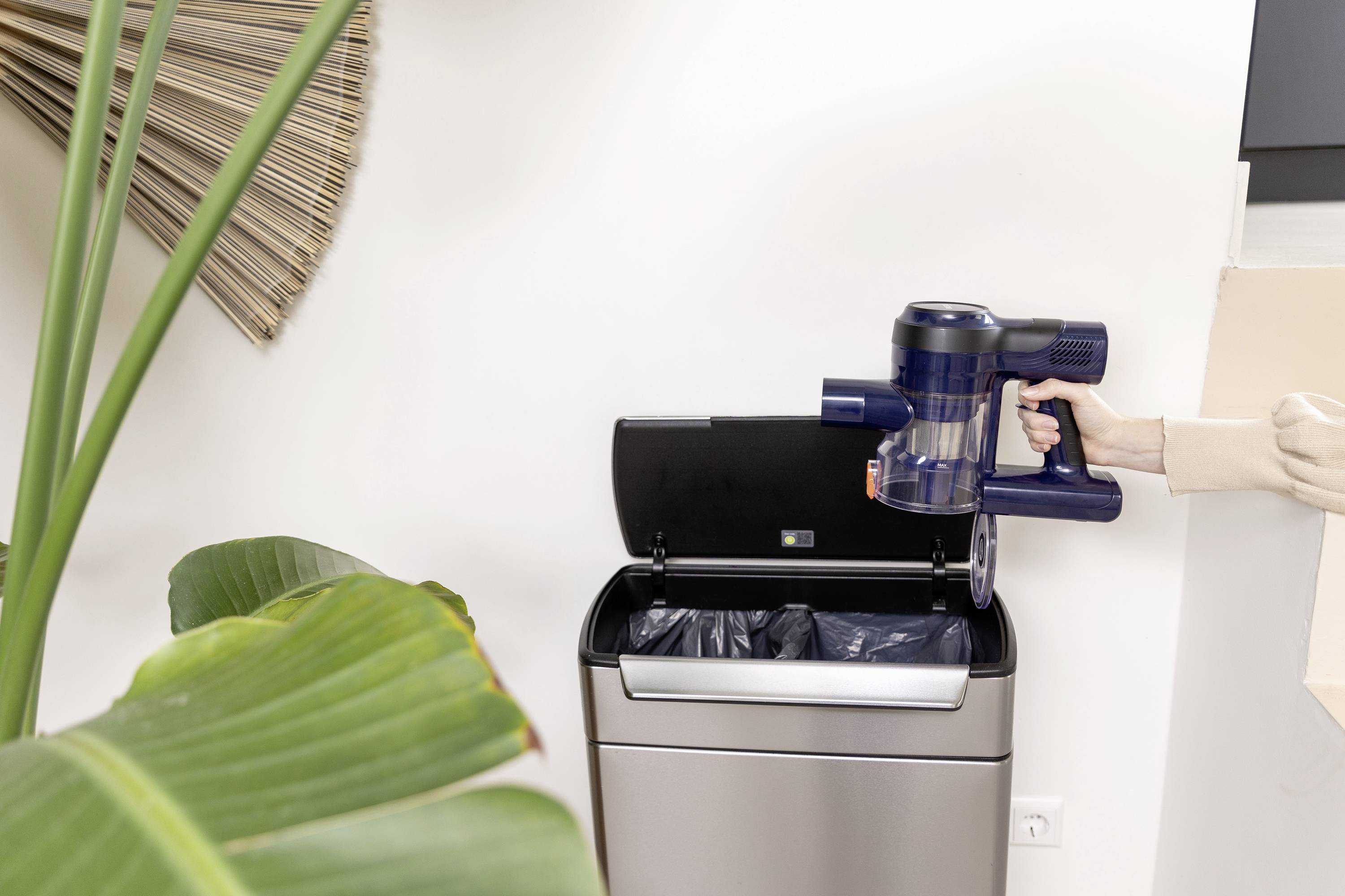 Une personne vide le réservoir à poussière d'un aspirateur sans fil dans une poubelle. Une plante d'intérieur et un décor moderne sont visibles en arrière-plan.