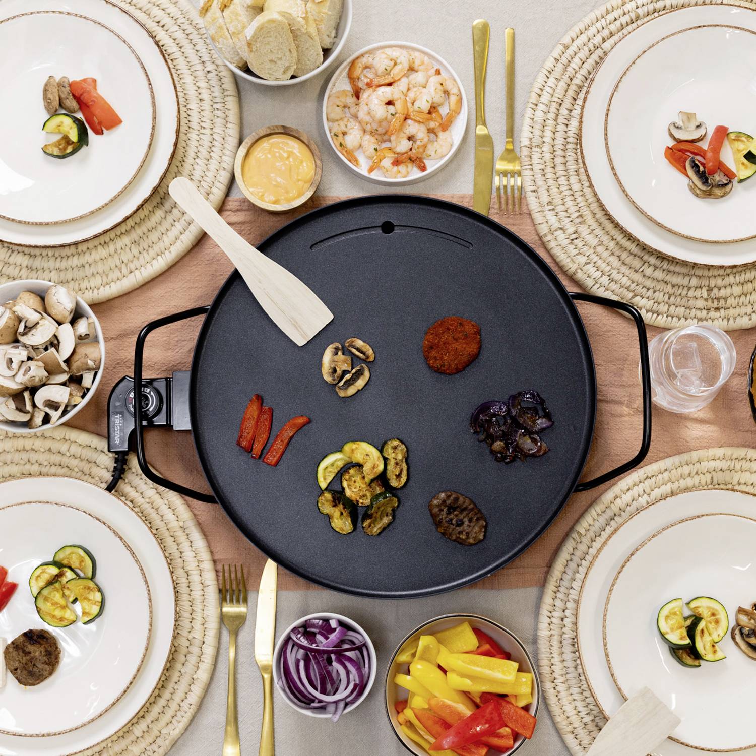 Un appareil de grill de table électrique au centre d'une table dressée avec des assiettes remplies de légumes, de crevettes et de pain dans des plats et des assiettes.