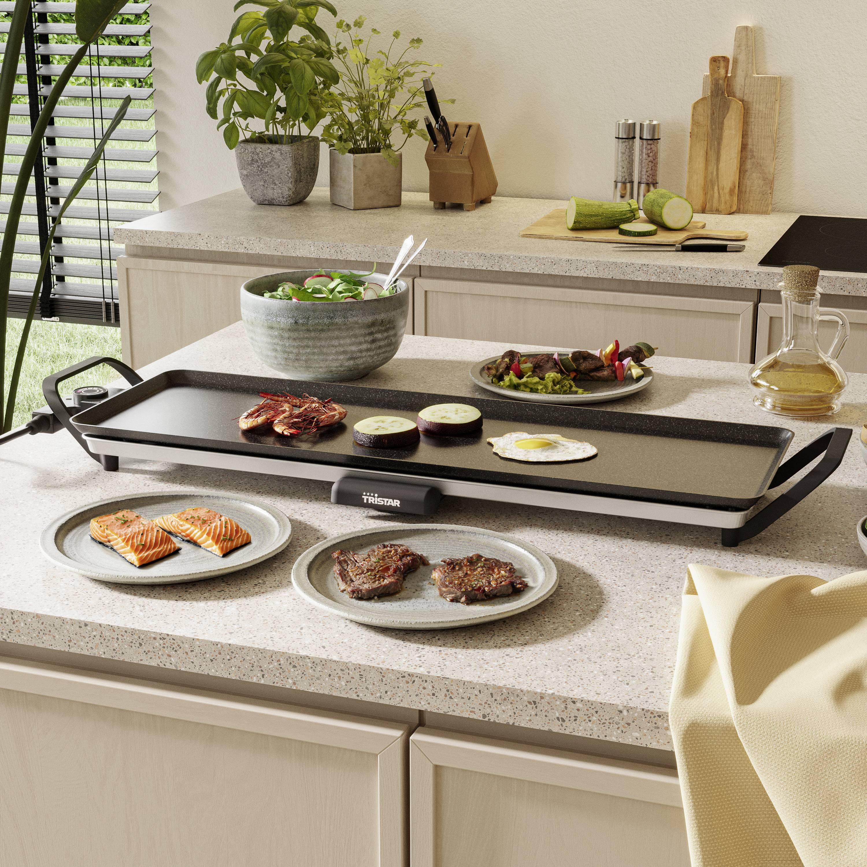Un grill de table sur un plan de travail de cuisine avec de la viande et des légumes grillés. Des ustensiles de cuisine et des plantes sont visibles en arrière-plan.