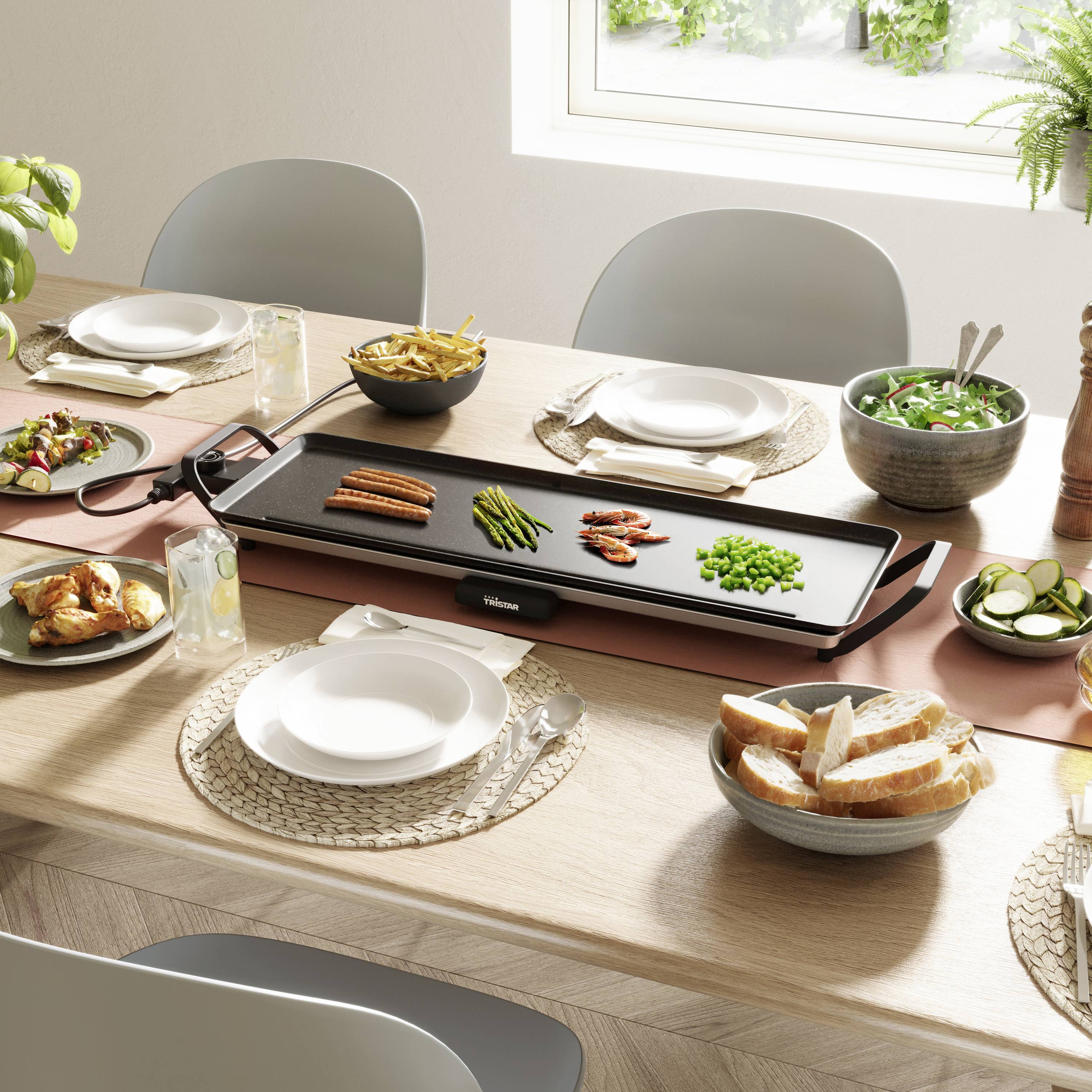 Une table dressée avec un grill électrique au centre, garnie de légumes et de viande, entourée de vaisselle et de pain.