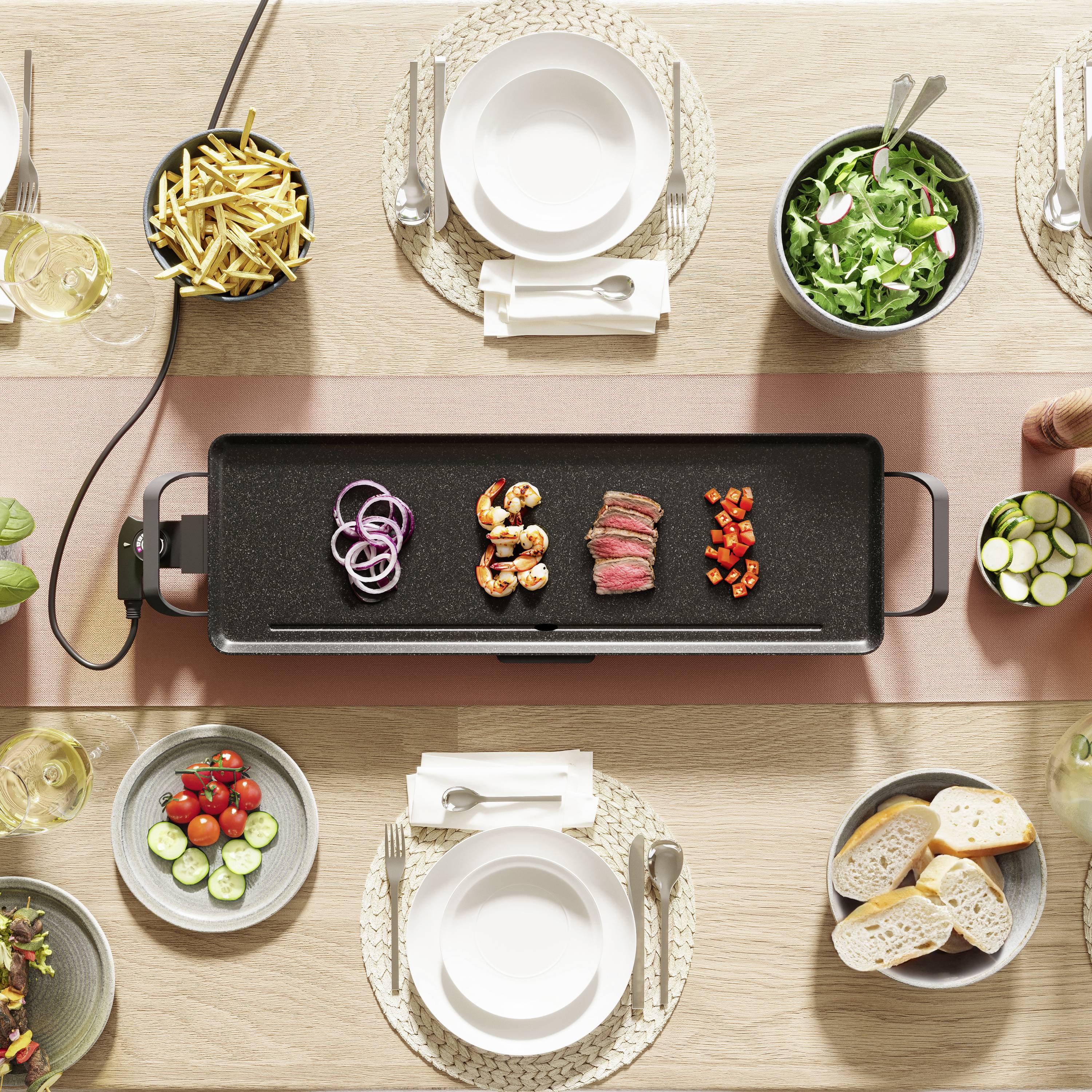 Une table avec une plaque de grill, sur laquelle se trouvent des crevettes, de la viande et des légumes. Tout autour, une salade, des frites, du pain et des assiettes vides avec des couverts.