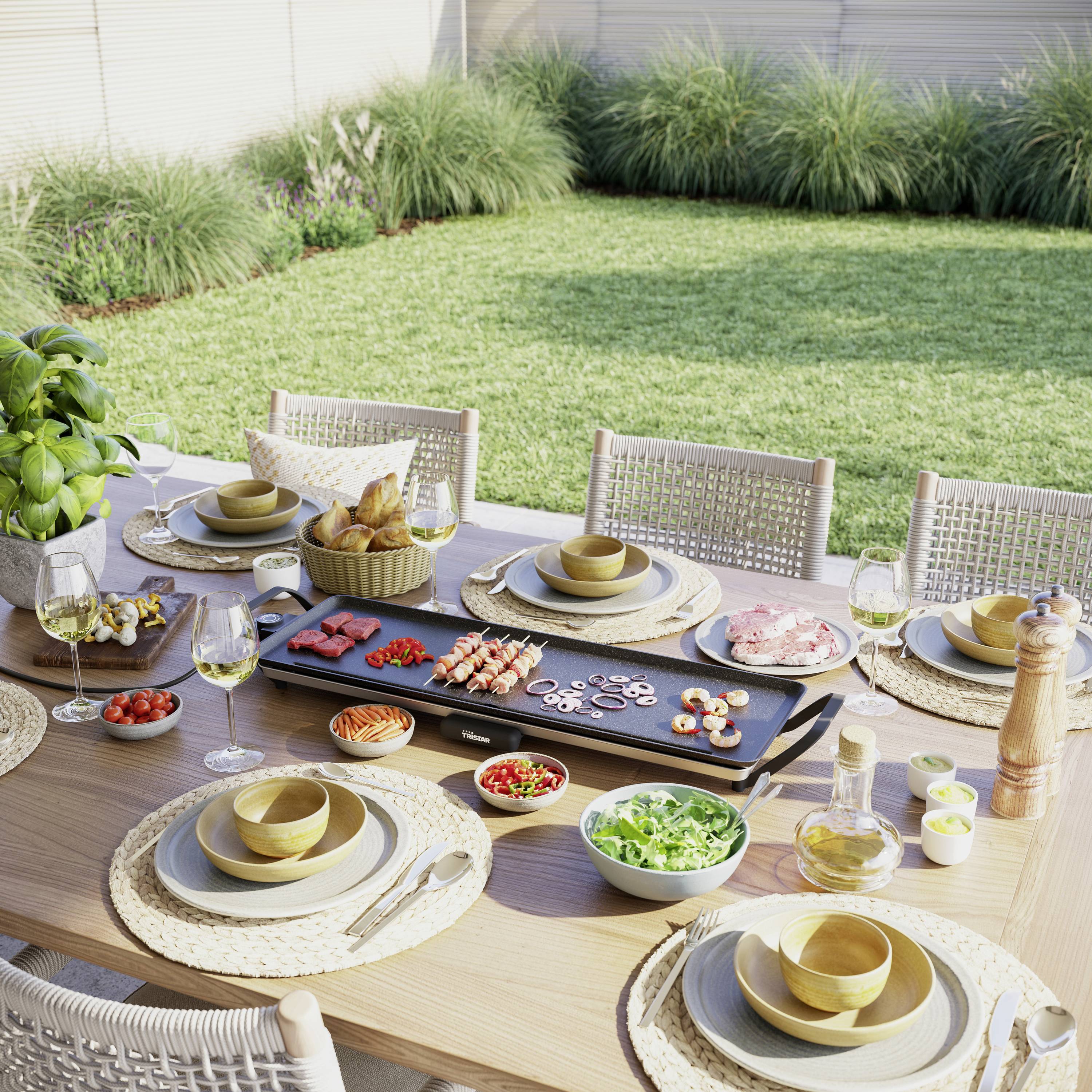 Une table dressée dans le jardin avec une plaque de grill, divers ingrédients, de la vaisselle et des boissons. À l'arrière-plan, pelouse et buissons.