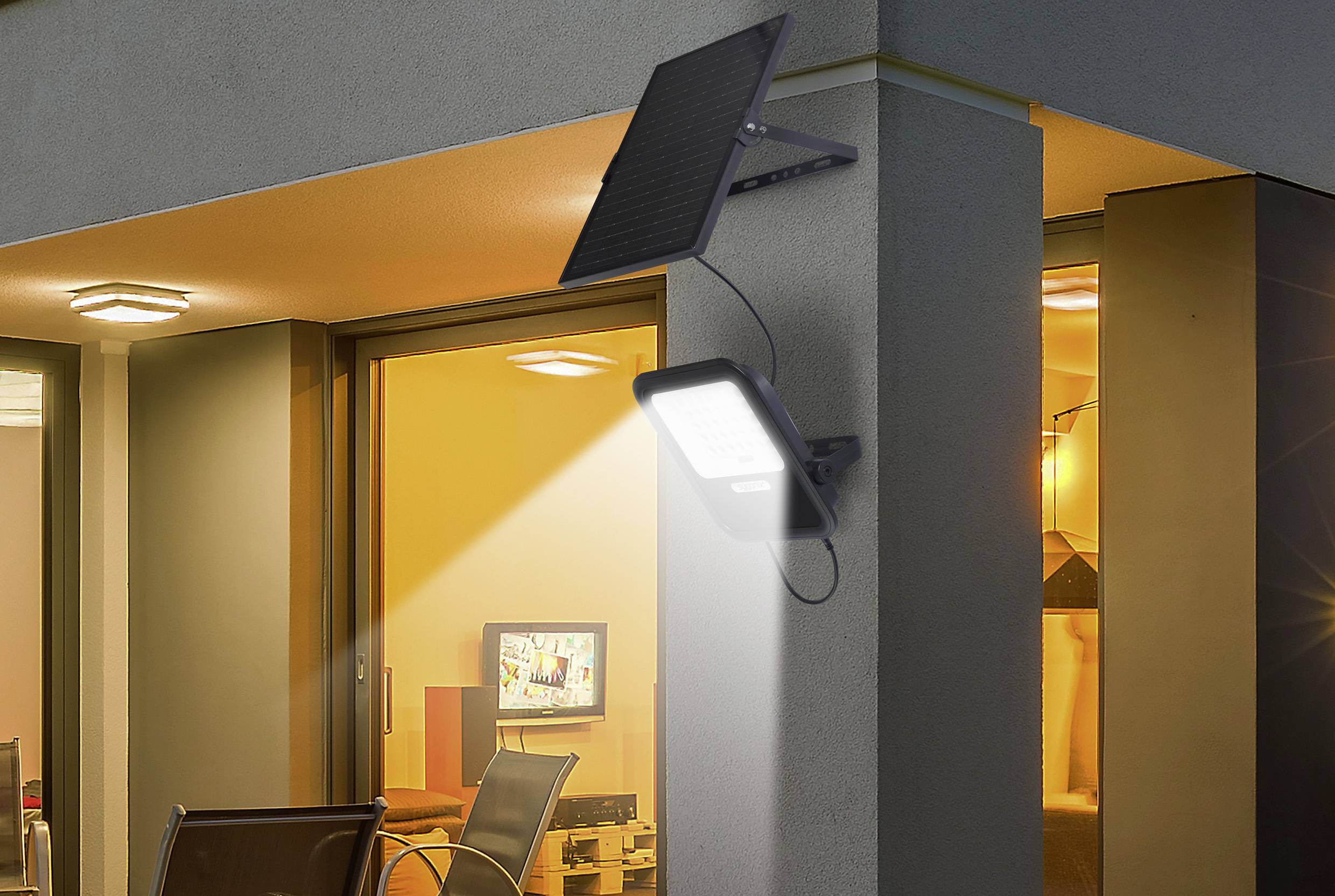Une lampe solaire est fixée à un mur de maison et éclaire la zone en dessous. À l'arrière-plan, on aperçoit une pièce illuminée.