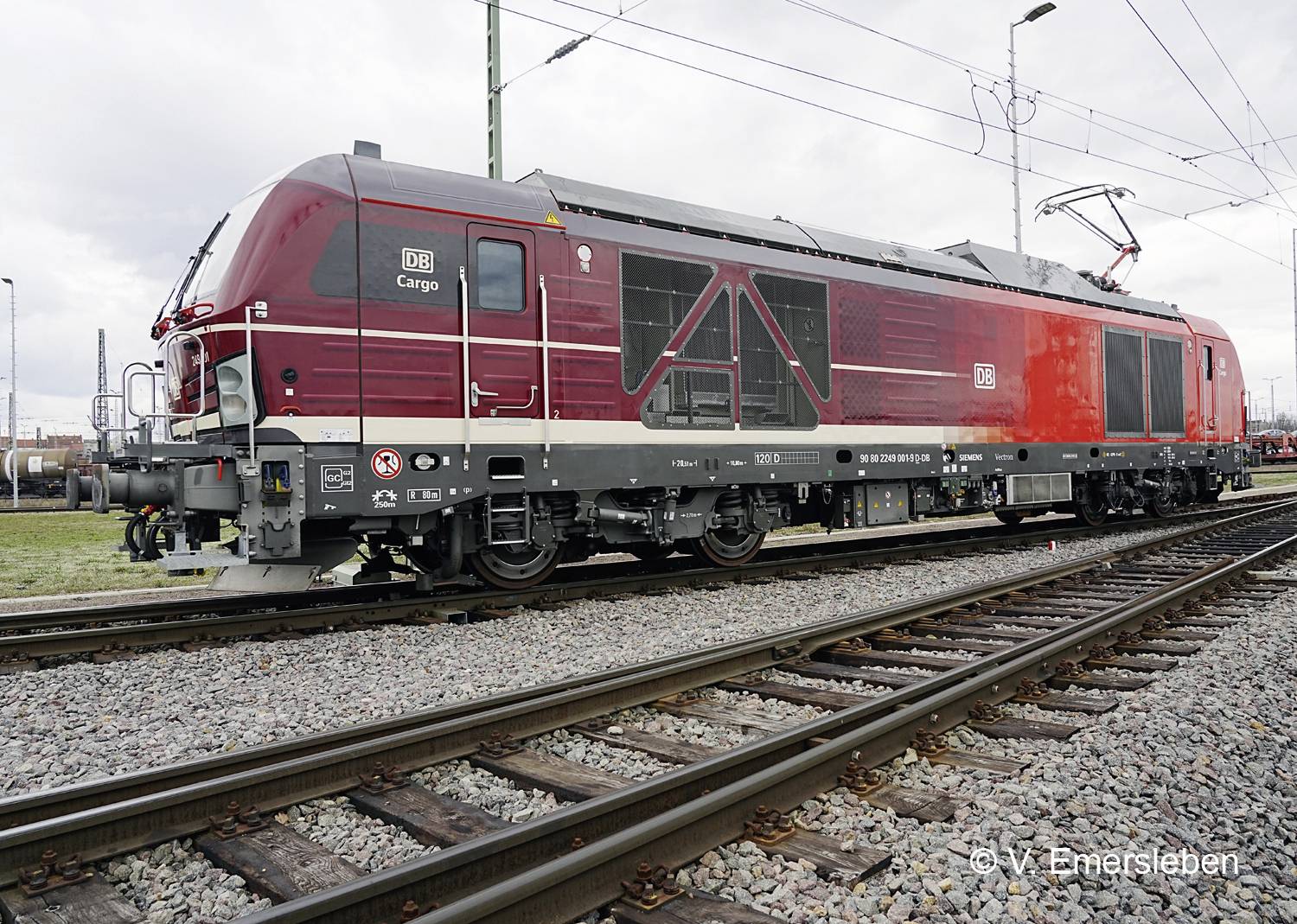 Une locomotive rouge et grise de DB Cargo sur des voies ferrées, vue latérale, sous un ciel nuageux.