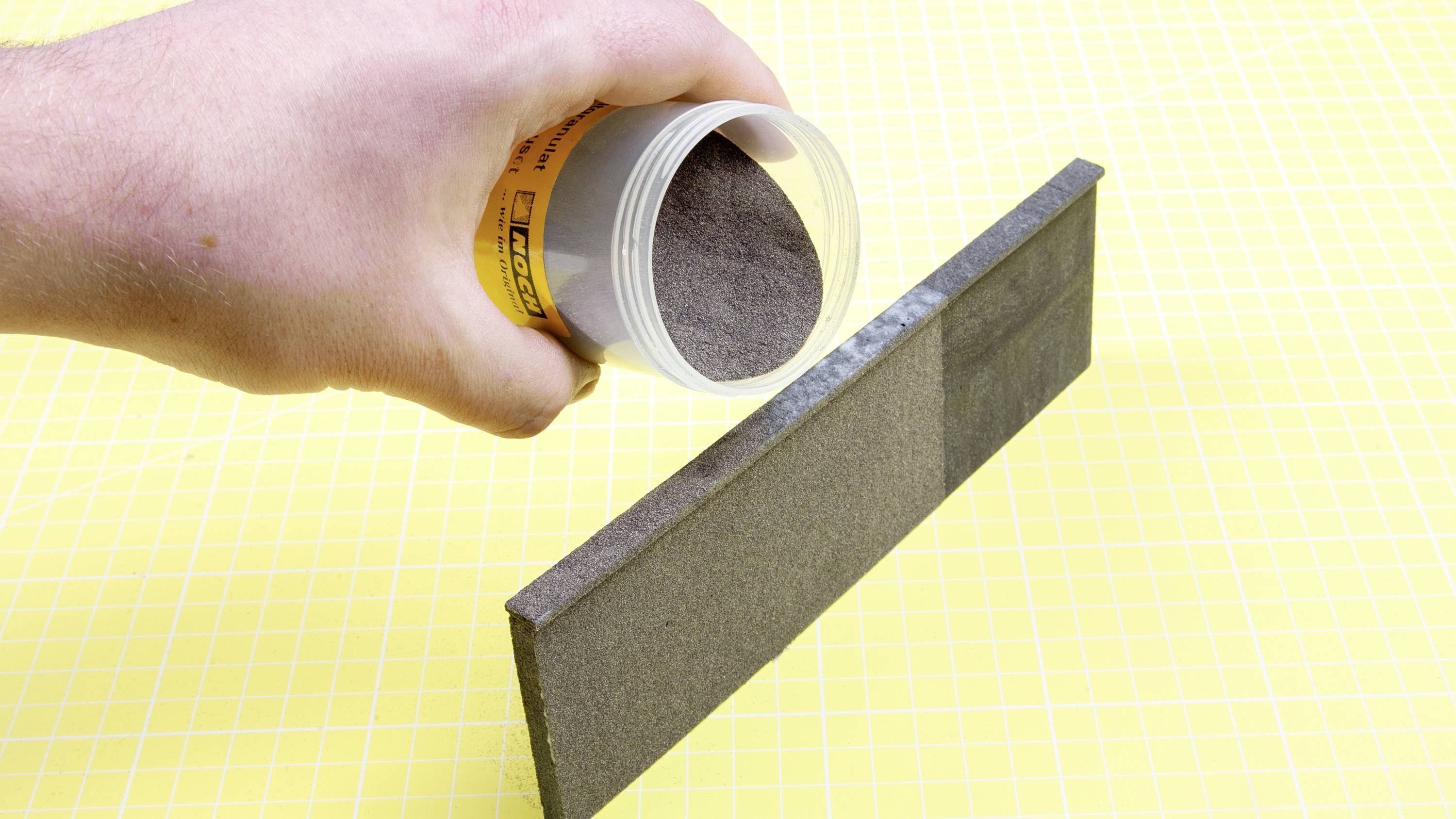 Une main verse une poudre grise d'un récipient sur une surface fine et rectangulaire qui est verticale. Fond jaune.
