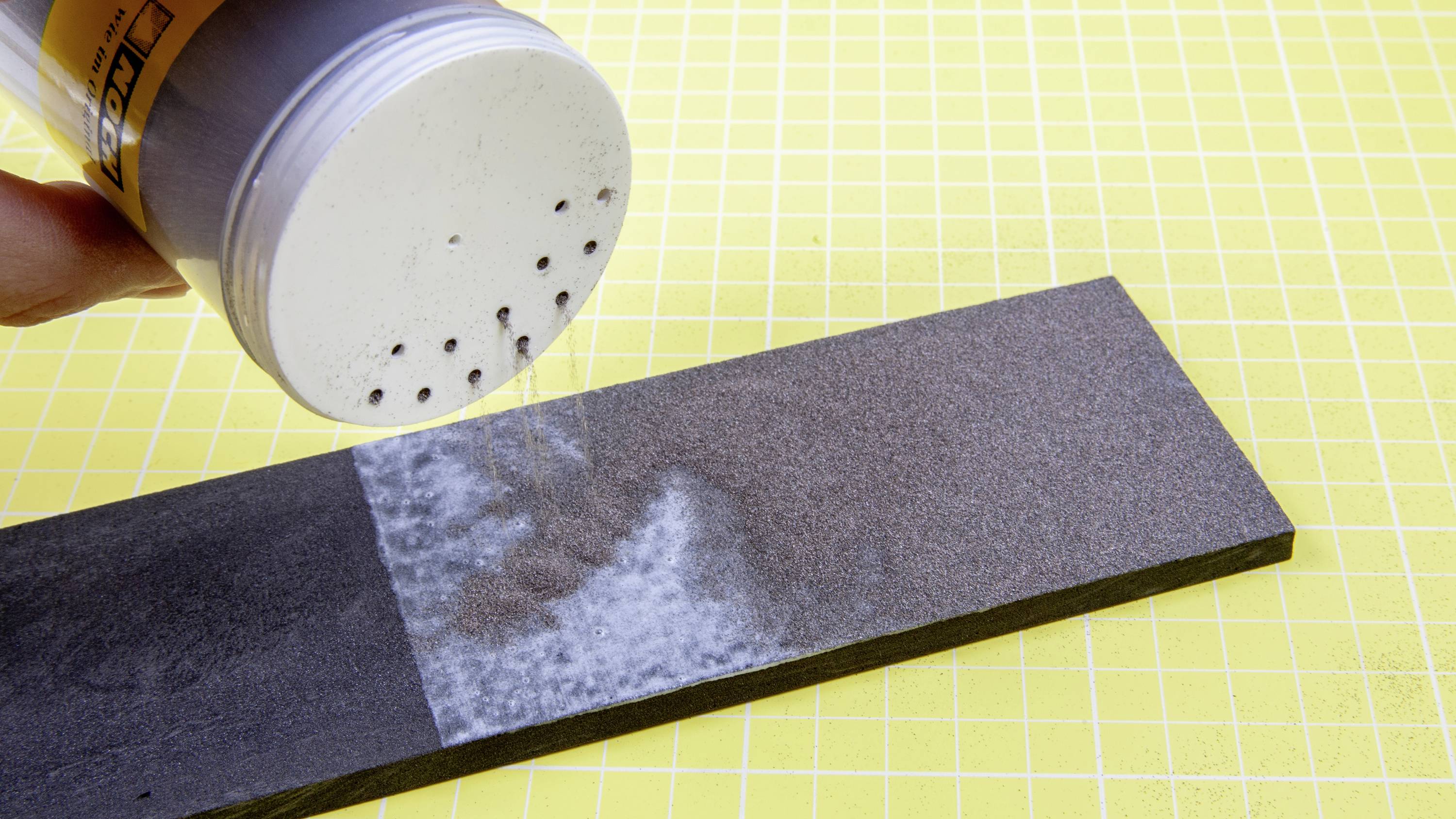 Une main répand de la poudre sur un bloc de papier de verre sur un fond jaune à carreaux. Une moitié du bloc montre la poudre, l'autre non.