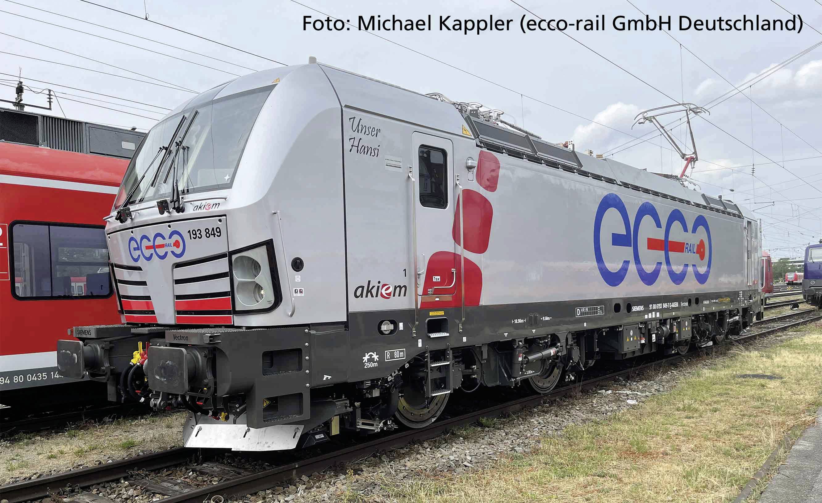 Une locomotive ECCO gris-argenté est stationnée à côté d'un train rouge sur une voie ferrée. Texte sur la locomotive : « Notre Hans ».