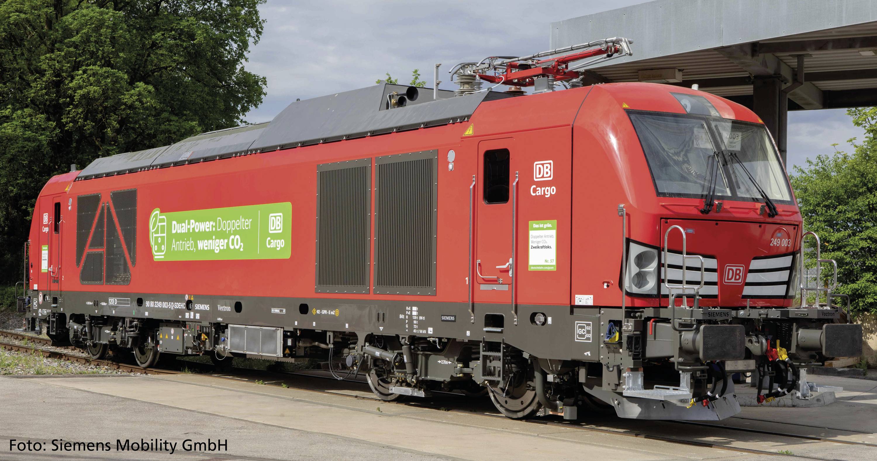 Train de marchandises rouge de DB Cargo stationné sur une voie. Inscription « Dual-Power : Double motorisation, moins de CO₂ ». Devant un quai couvert.