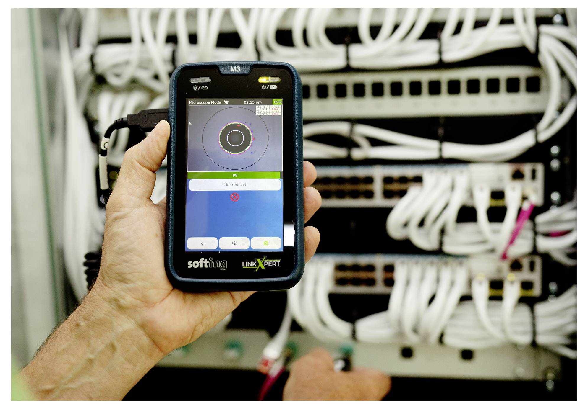 Hand, die ein tragbares Netzwerktestgerät vor einem Serverrack mit vielen verbundenen Ethernet-Kabeln und Schaltern hält.