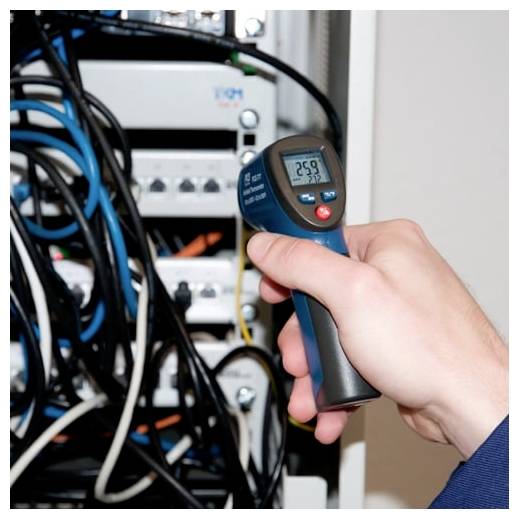 Main tenant un thermomètre infrarouge numérique pointé vers un équipement électrique et des câbles emmêlés à l'intérieur d'une armoire.