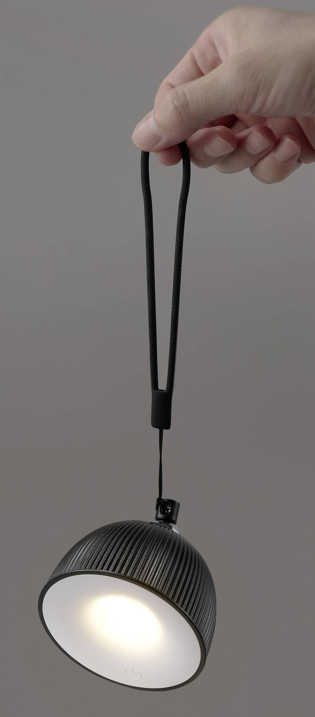 Une main tient une petite lampe suspendue noire avec une poignée flexible, illuminée sur un fond gris.