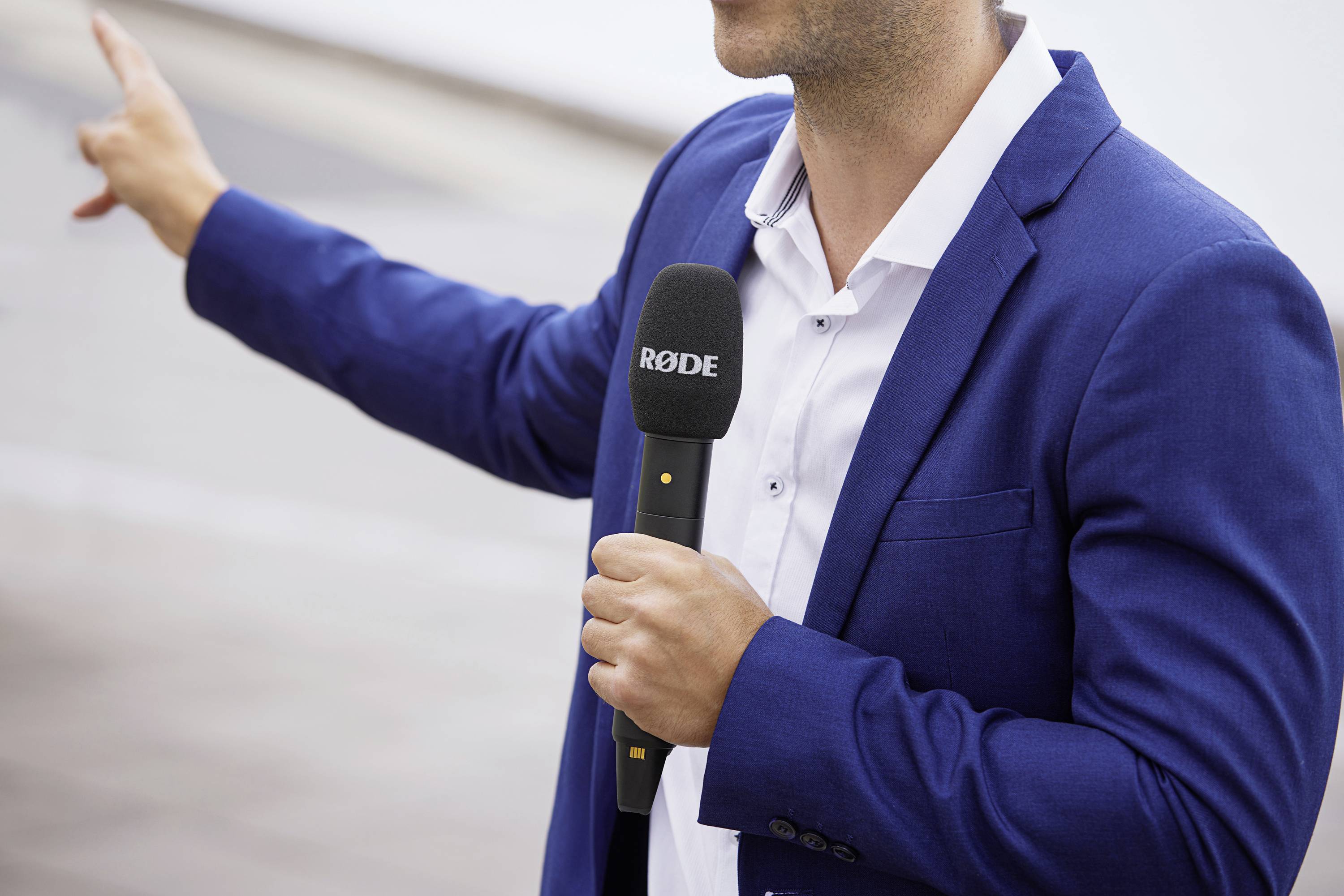 Une personne portant un costume bleu tient un microphone, pointe vers quelque chose en dehors de l'image et semble parler, probablement lors d'une interview.