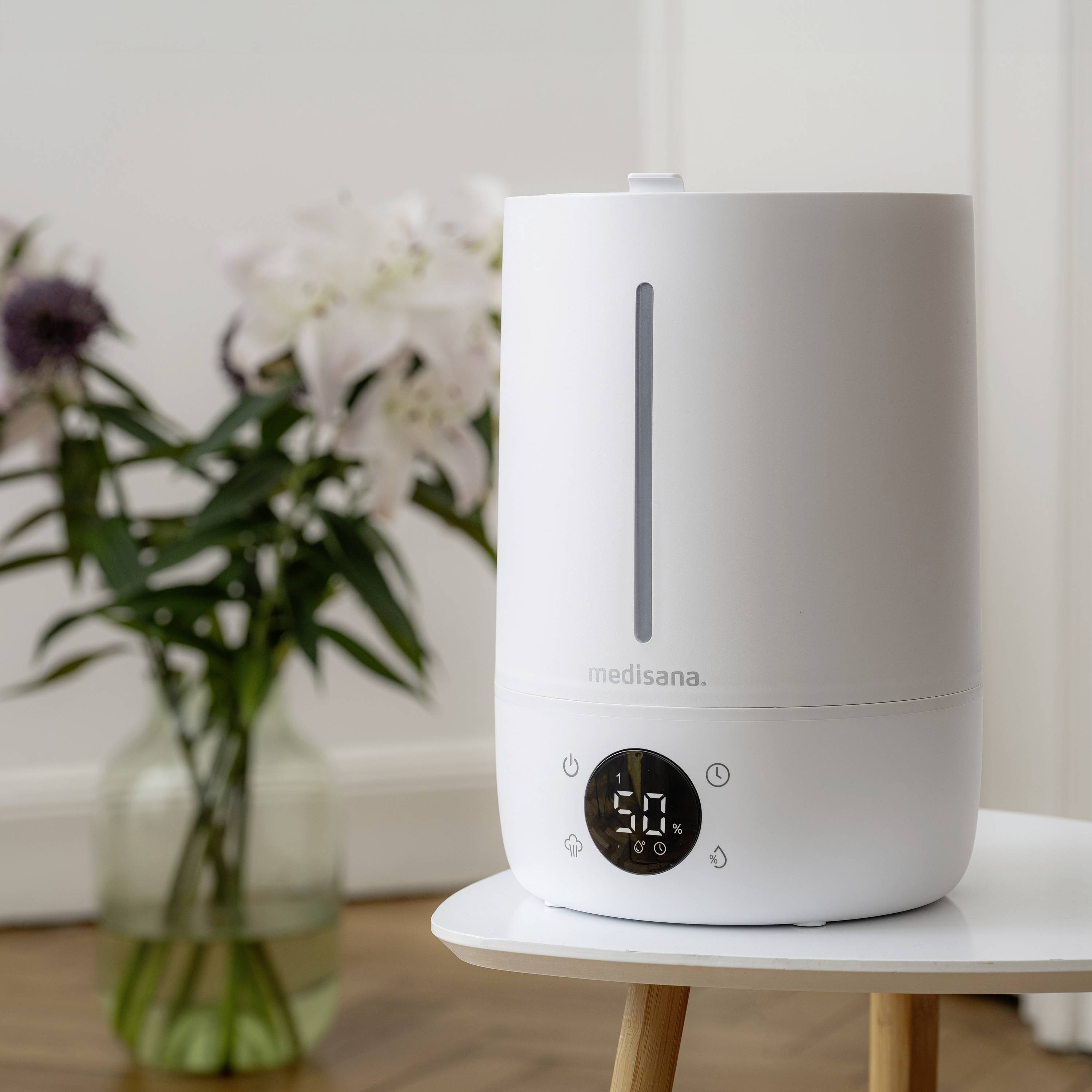 Un humidificateur blanc est posé sur une table devant un arrière-plan doux. En arrière-plan, des fleurs floues sont placées dans un vase.