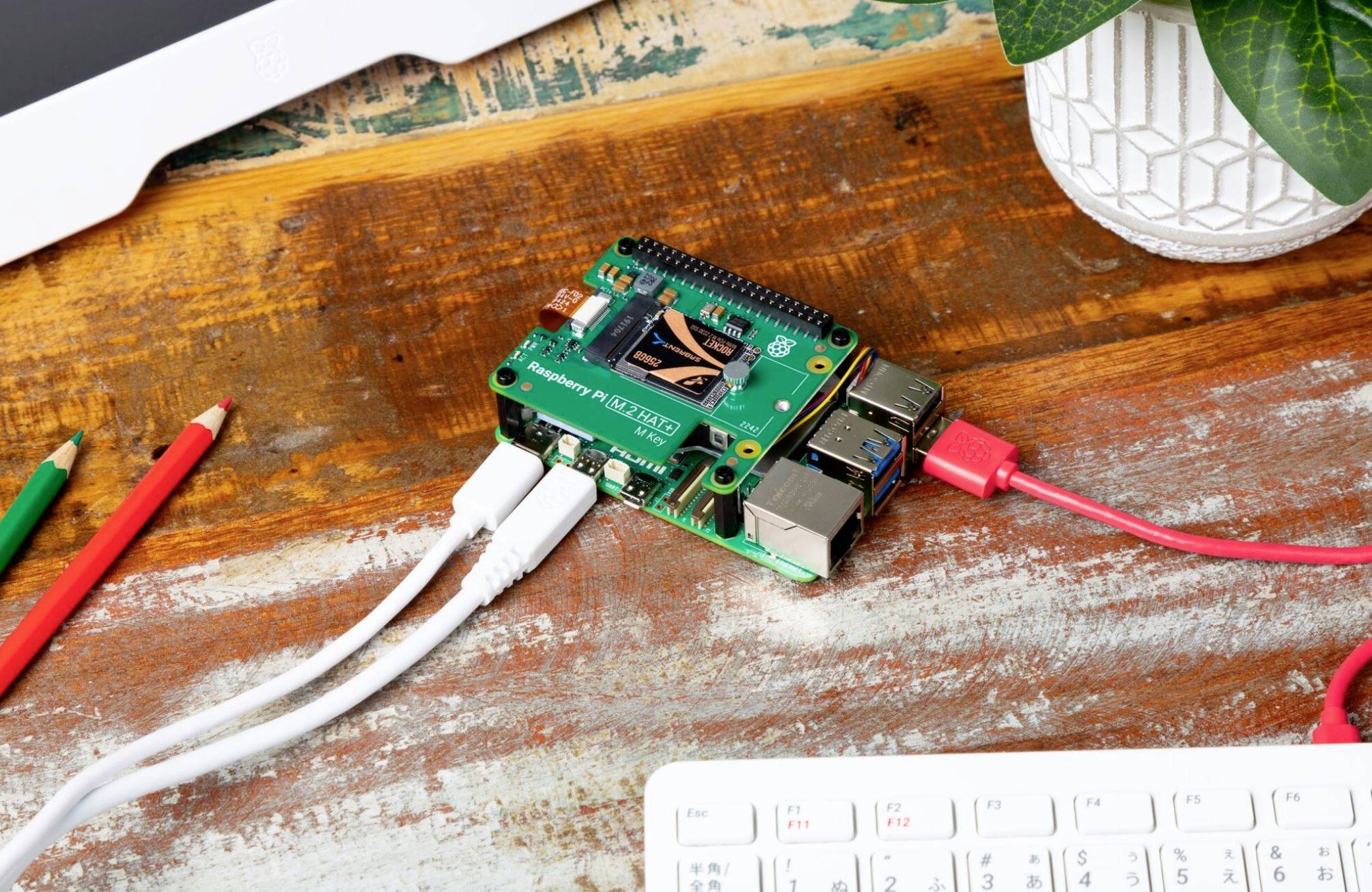 Sur une table, un Raspberry Pi avec des câbles connectés est posé à côté de crayons et d'une plante, entouré d'équipement informatique.