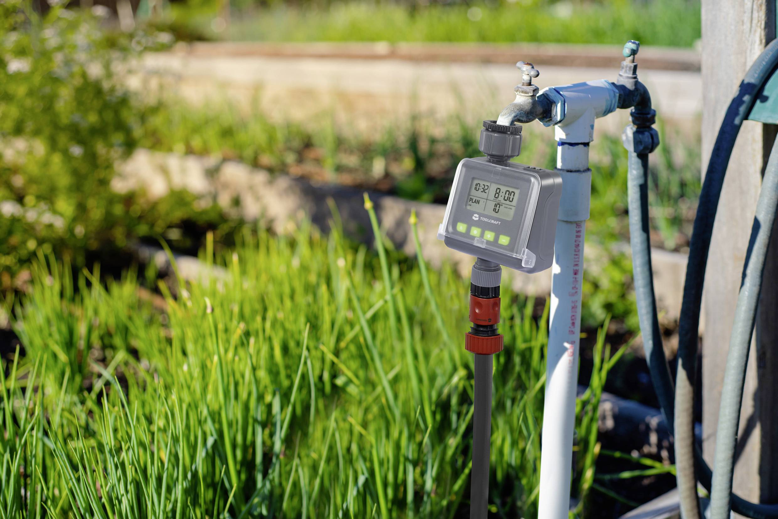 Un minuteur d'irrigation numérique est connecté à un tuyau d'arrosage et se trouve dans un jardin avec de l'herbe verte en arrière-plan.
