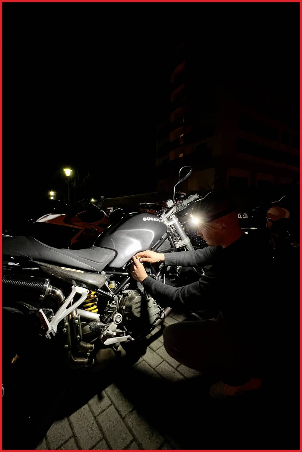 Une personne travaille la nuit sur une moto sur un trottoir sombre. Une lampe de poche éclaire la scène et la moto.