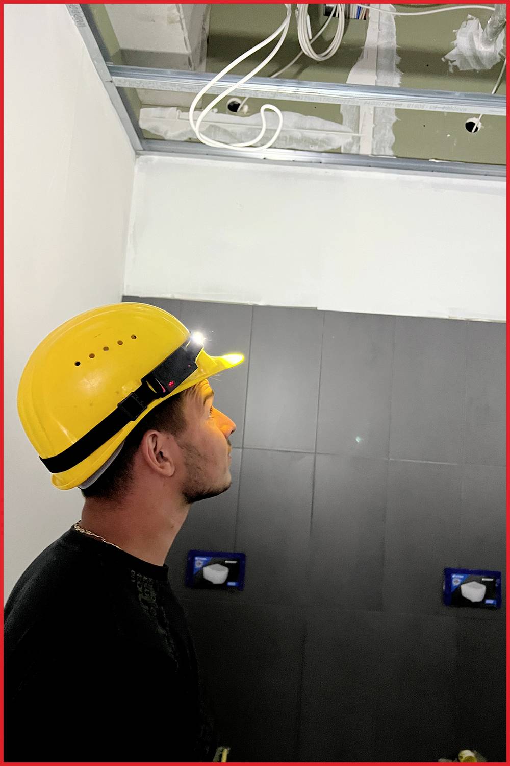 Un homme portant un casque jaune regarde vers le haut alors qu'il se trouve dans une pièce. Des tuyaux et des câbles sont présents au plafond.