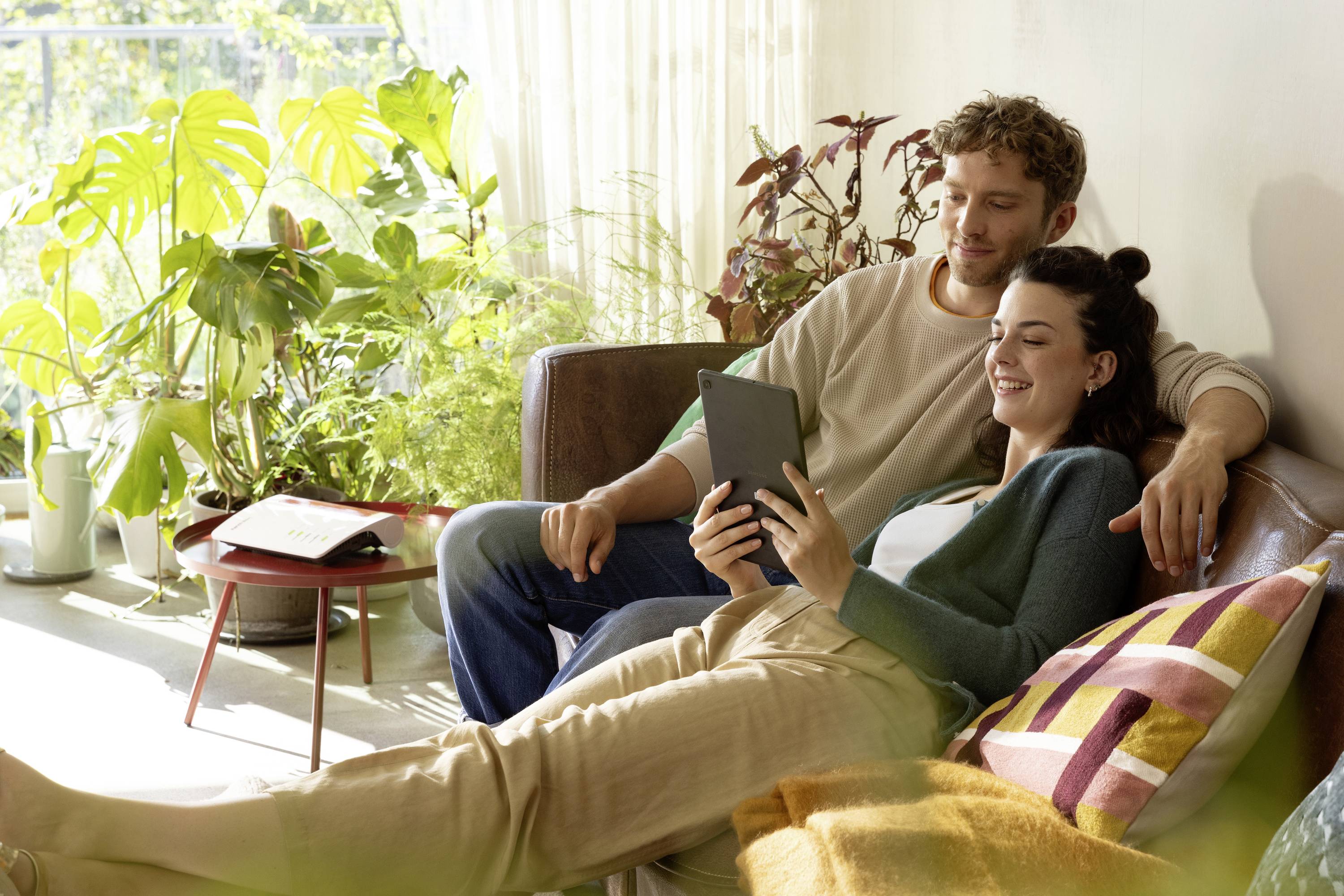 Un couple est assis sur un canapé et regarde ensemble une tablette. Ils sourient, entourés de plantes. Une lumière claire baigne la scène.