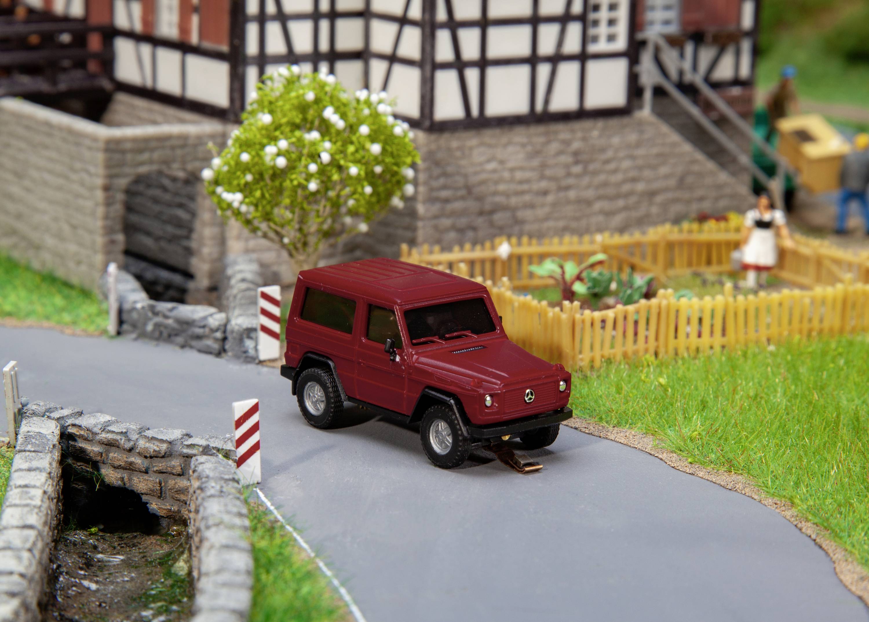 Une petite voiture miniature rouge est garée sur une route de campagne devant une maison à colombages, avec une clôture de jardin et un arbre en arrière-plan.