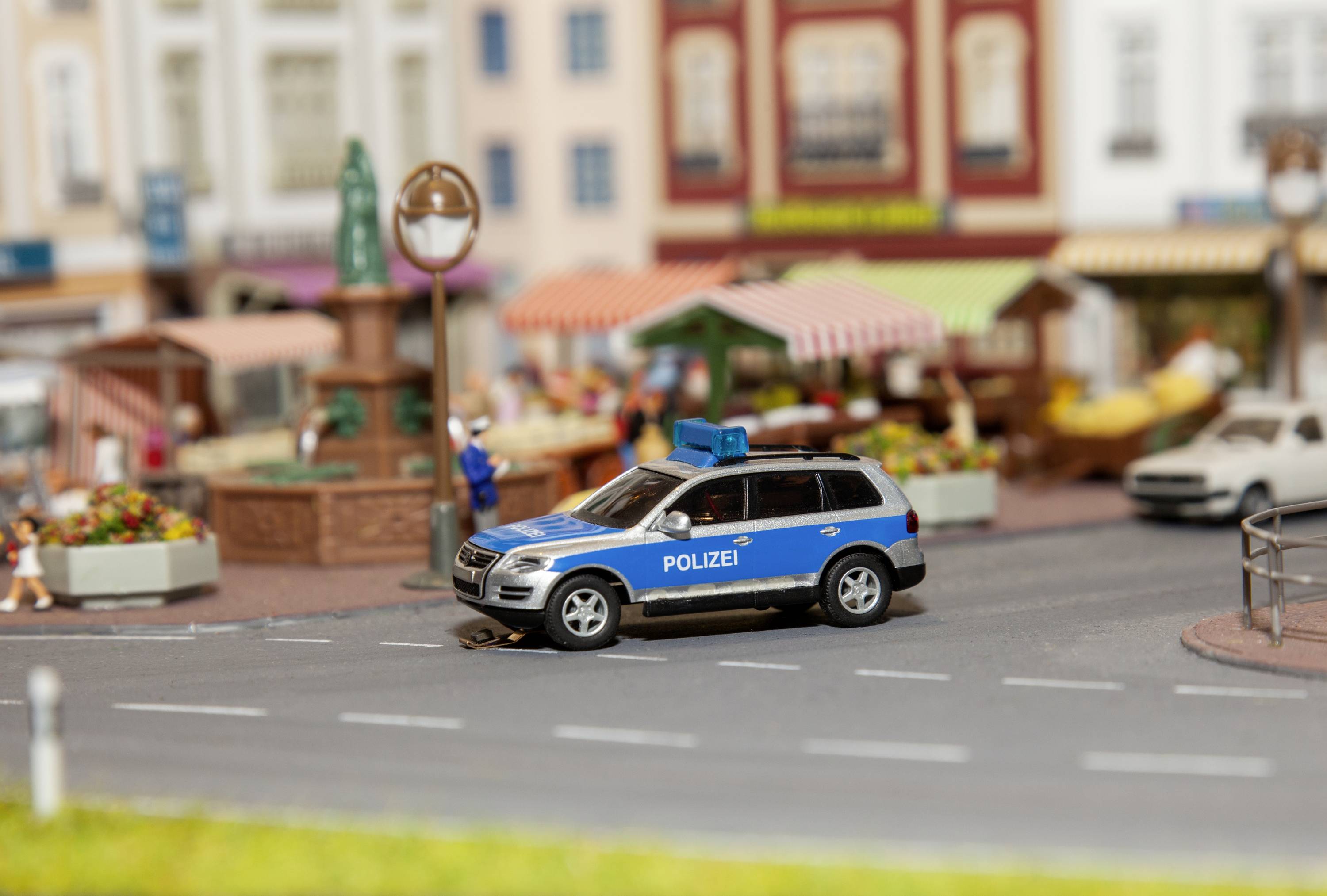 Une voiture de police miniature dans le style d'un véhicule allemand est garée sur une route miniature devant une place de marché modélisée avec des stands.