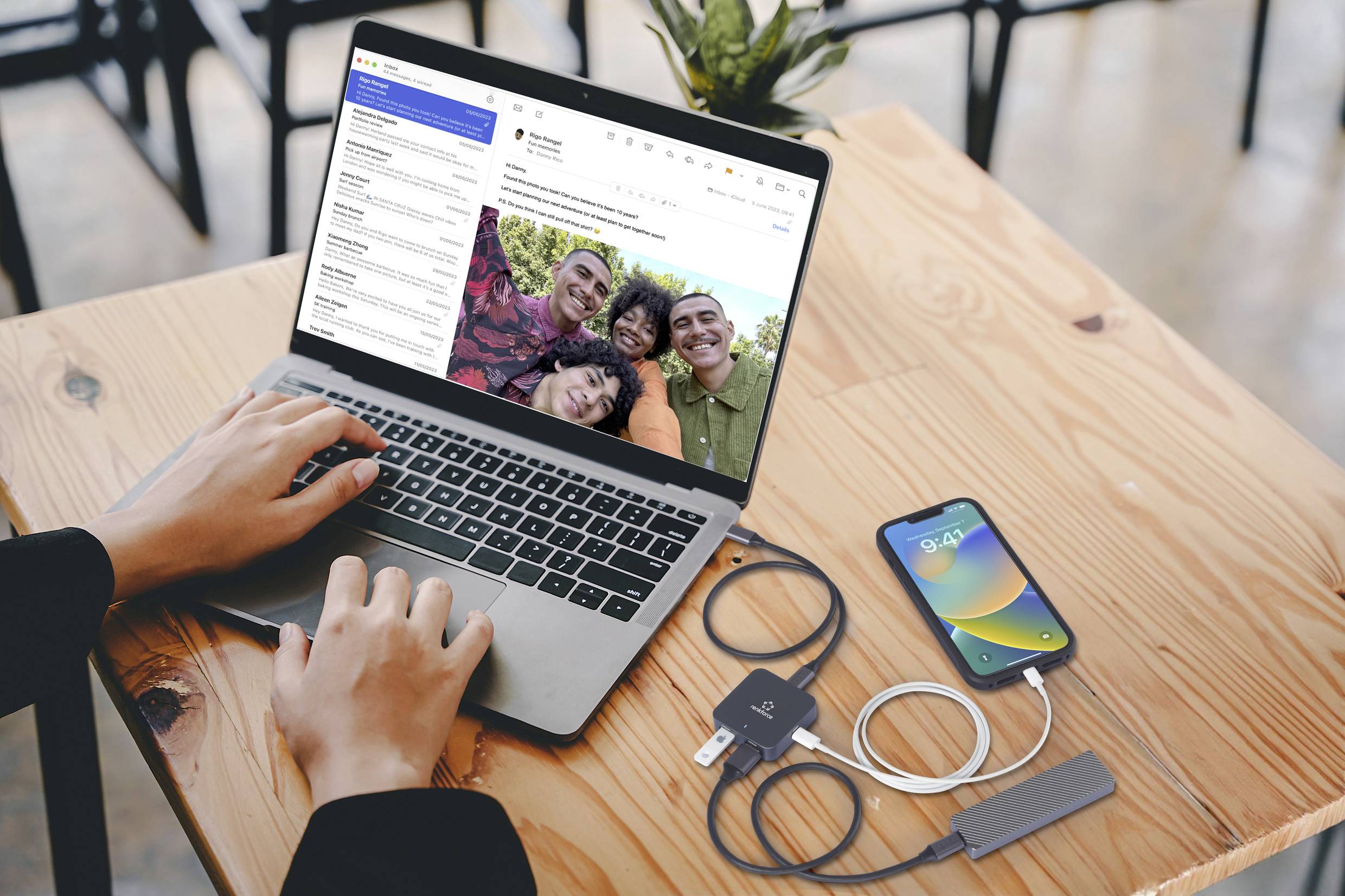 Une personne tape sur un ordinateur portable avec une boîte de réception e-mail ouverte, à côté se trouvent un smartphone avec un câble de chargement et d'autres câbles sur une table en bois.