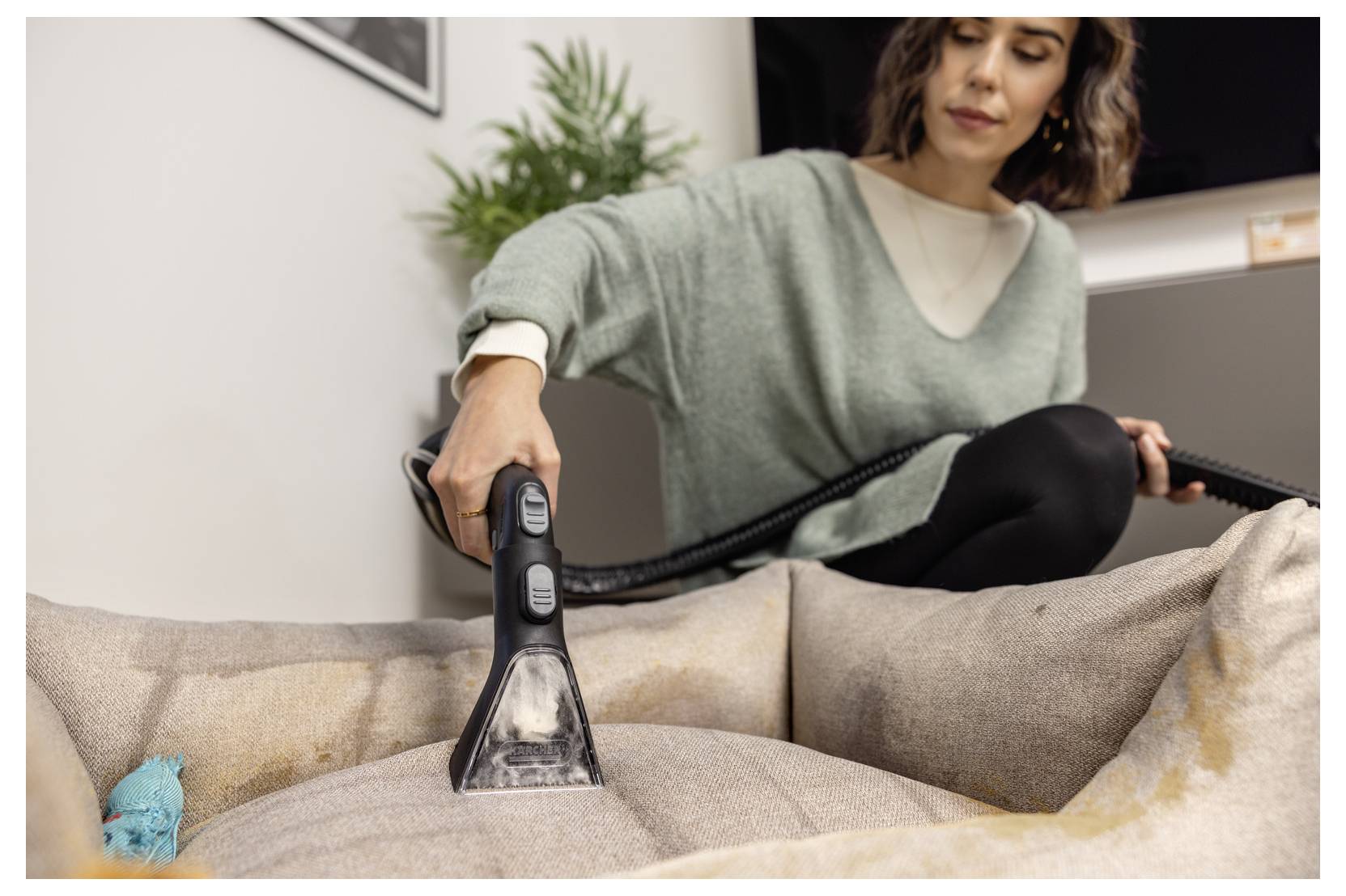 Une personne utilise un aspirateur portable avec une brosse d'accessoire pour nettoyer un canapé en tissu. Une petite plante est visible en arrière-plan.