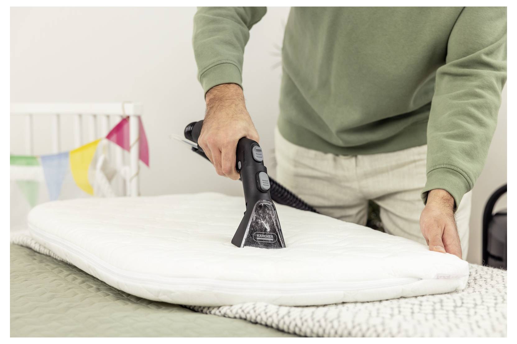 Une personne portant un pull vert nettoie à la vapeur un coussin blanc sur un lit, avec des drapeaux colorés suspendus en arrière-plan, suggérant un nettoyage domestique.