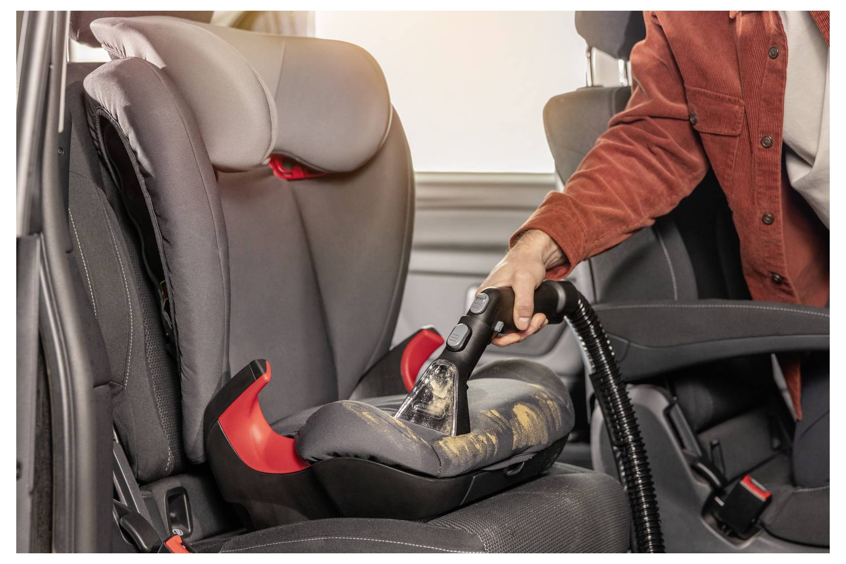 Une personne portant une veste rouge nettoie un siège auto pour enfant avec un aspirateur, éliminant les miettes et les débris à l'intérieur d'une voiture.