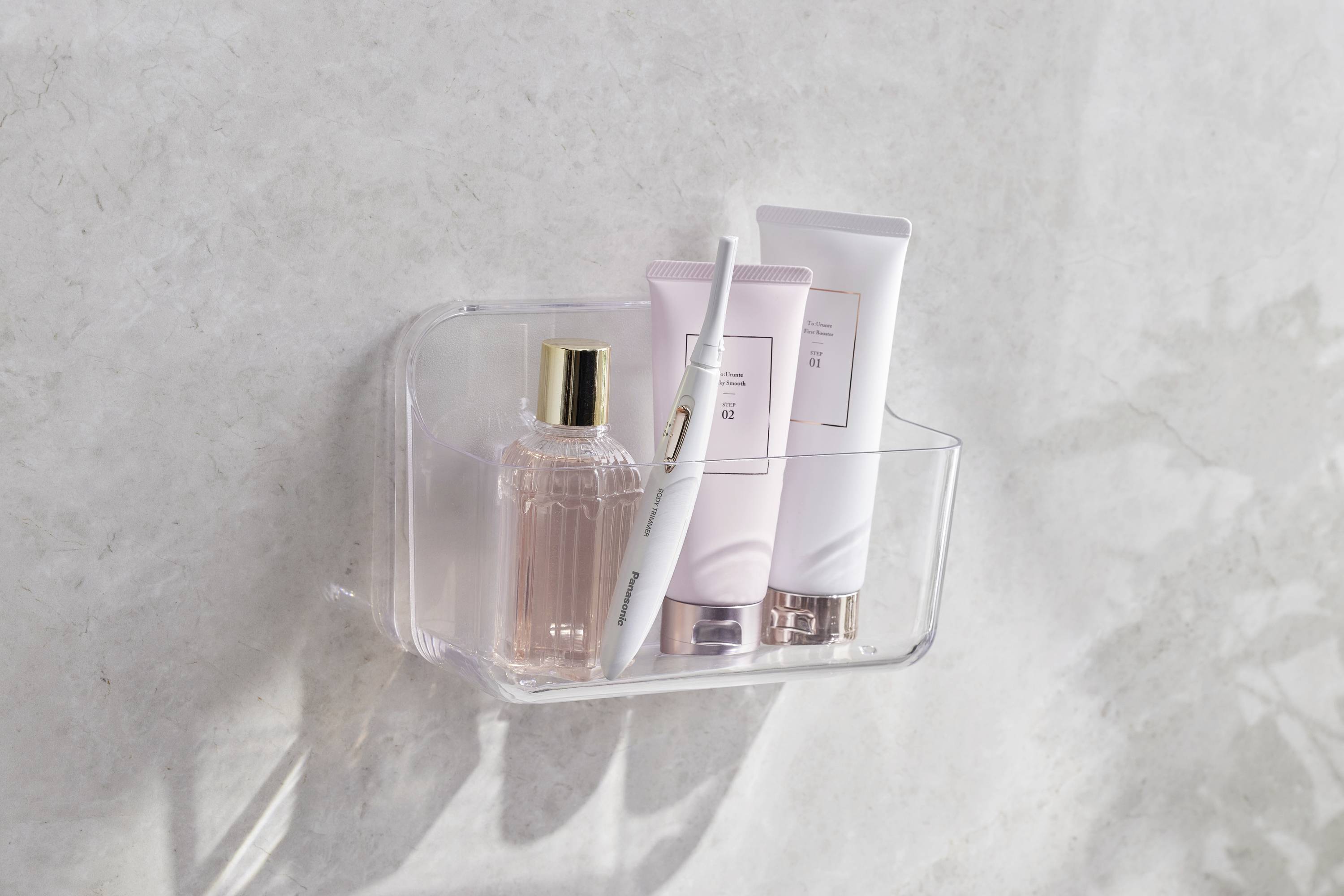 Un support transparent fixé au mur contient un flacon de parfum, deux tubes et une brosse à dents électrique.