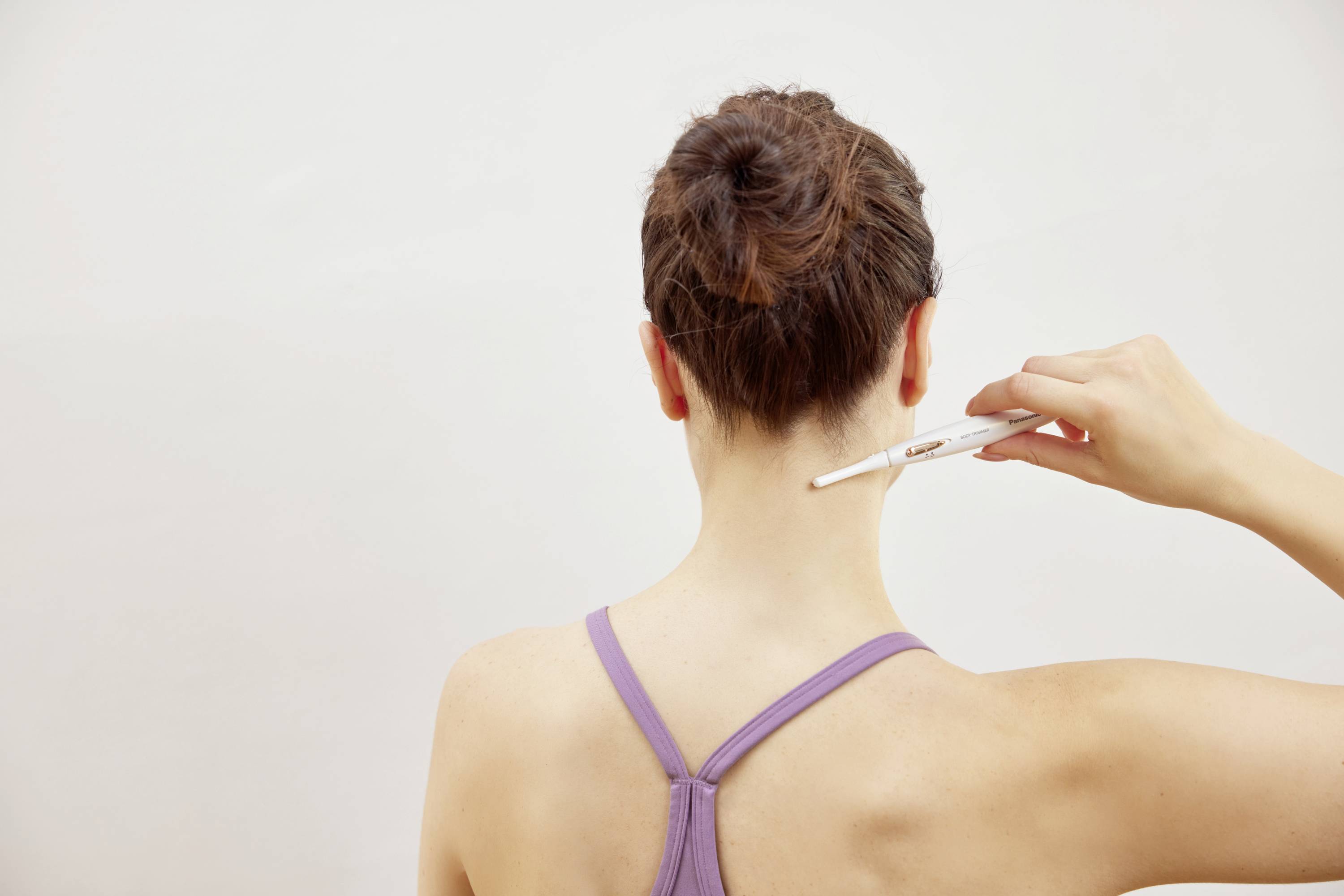 Une personne pointe son propre nuque avec un stylo blanc. Elle porte un haut lilas. L'arrière-plan est gris et neutre.