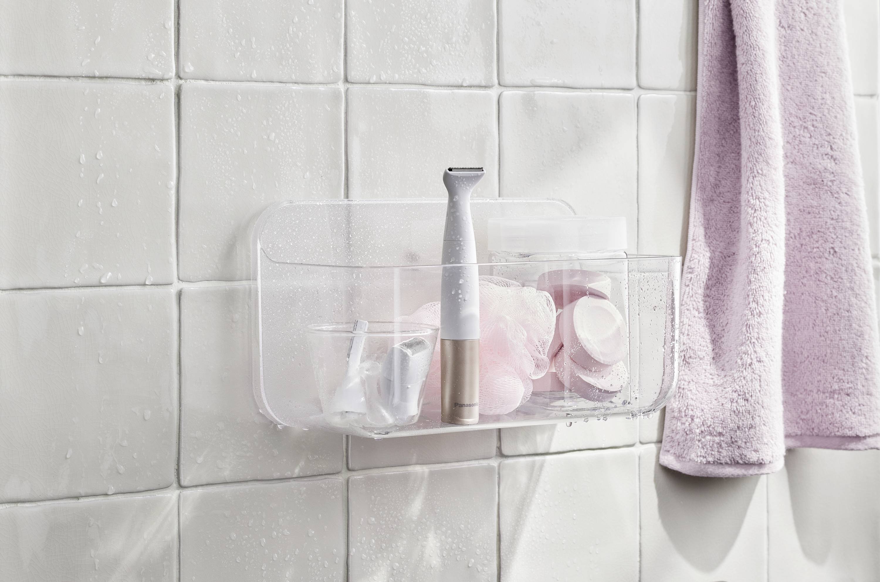 Étagère de douche avec des articles d'hygiène et un rasoir électrique, à côté d'une serviette rose. Carreaux blancs en arrière-plan.