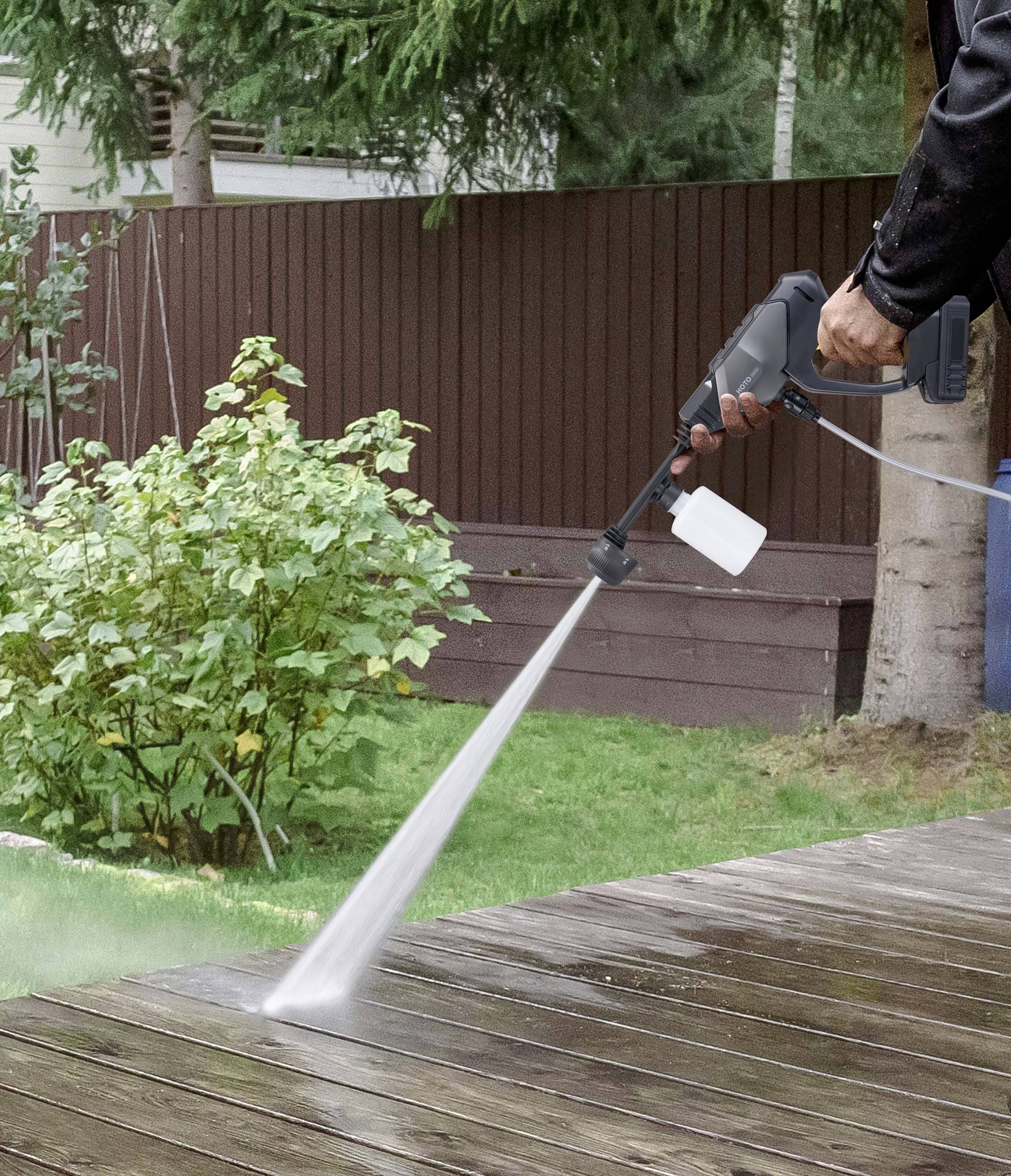 Une personne nettoie une terrasse en bois à l'aide d'un nettoyeur haute pression ; des arbustes et une clôture sont visibles en arrière-plan.