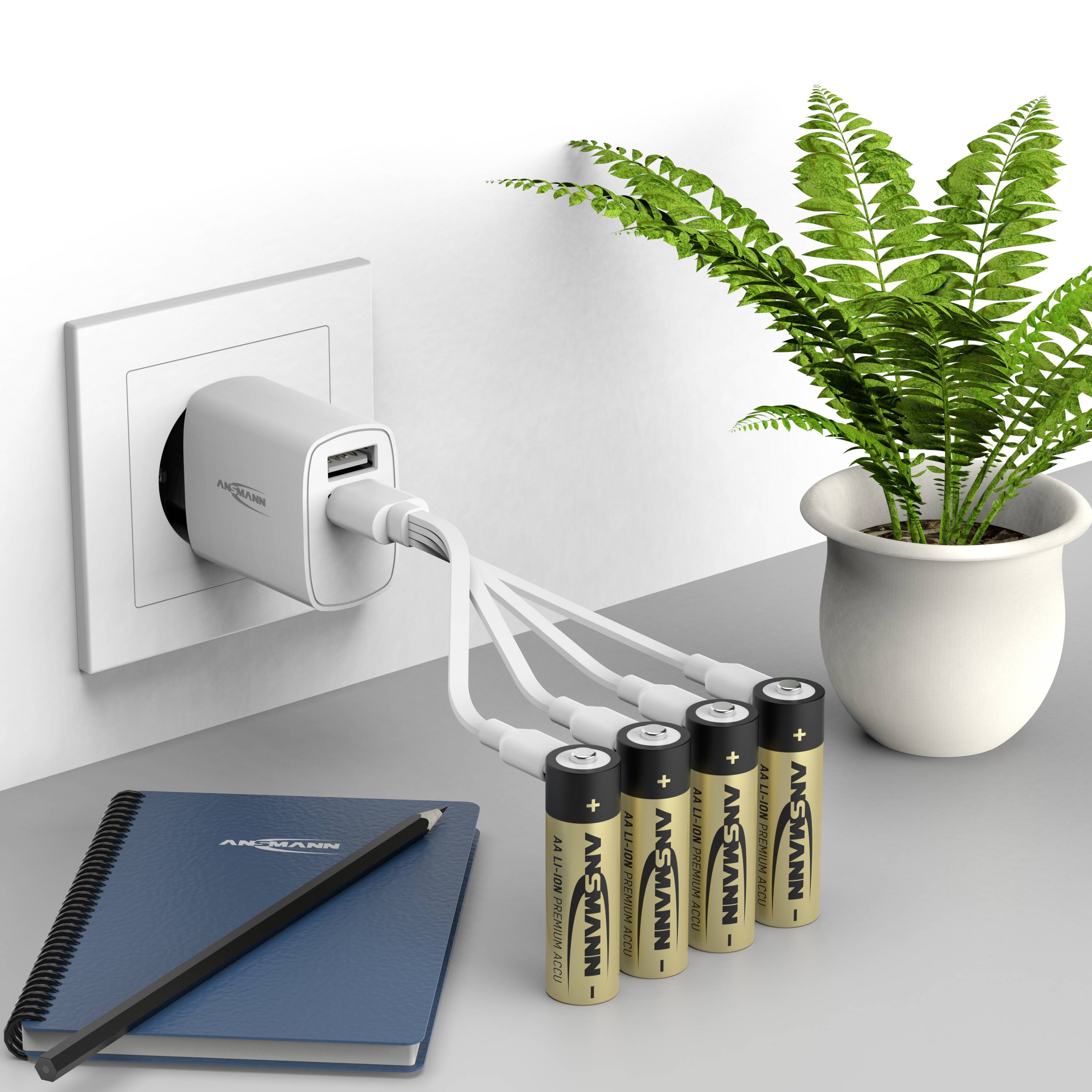 Une prise électrique avec un adaptateur de charge USB charge quatre piles AA. À côté, une plante verte dans un pot et un carnet sur la table.