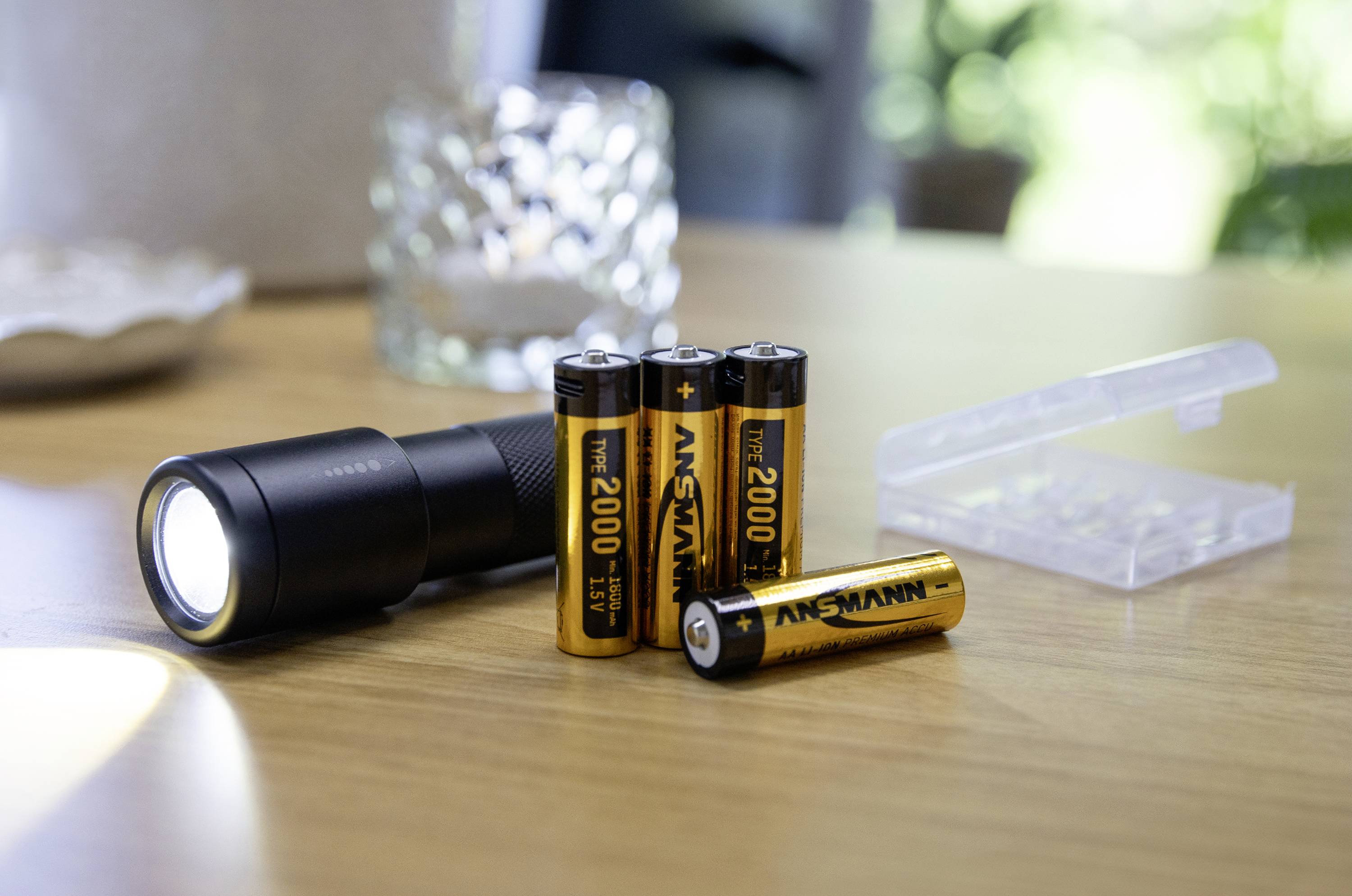 Une lampe de poche noire brille sur une table en bois à côté de trois piles AA et d'un étui transparent pour batteries.
