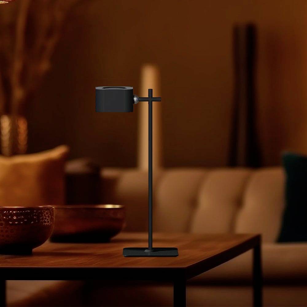 Une lampe de table moderne avec un abat-jour orientable est posée sur une table en bois dans une pièce doucement éclairée, avec un canapé flou en arrière-plan.