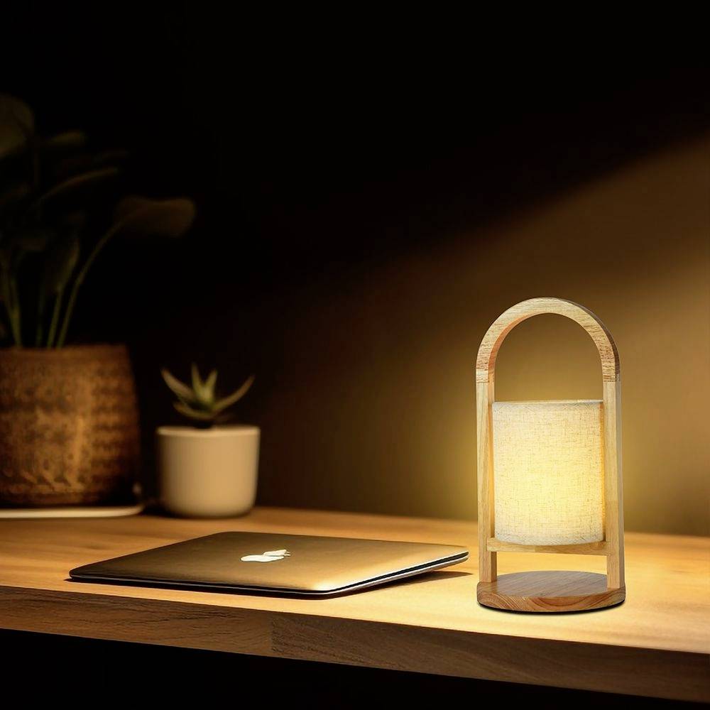 Un ordinateur portable est posé sur une table en bois à côté d'une lampe de bureau allumée, avec une plante visible en arrière-plan.