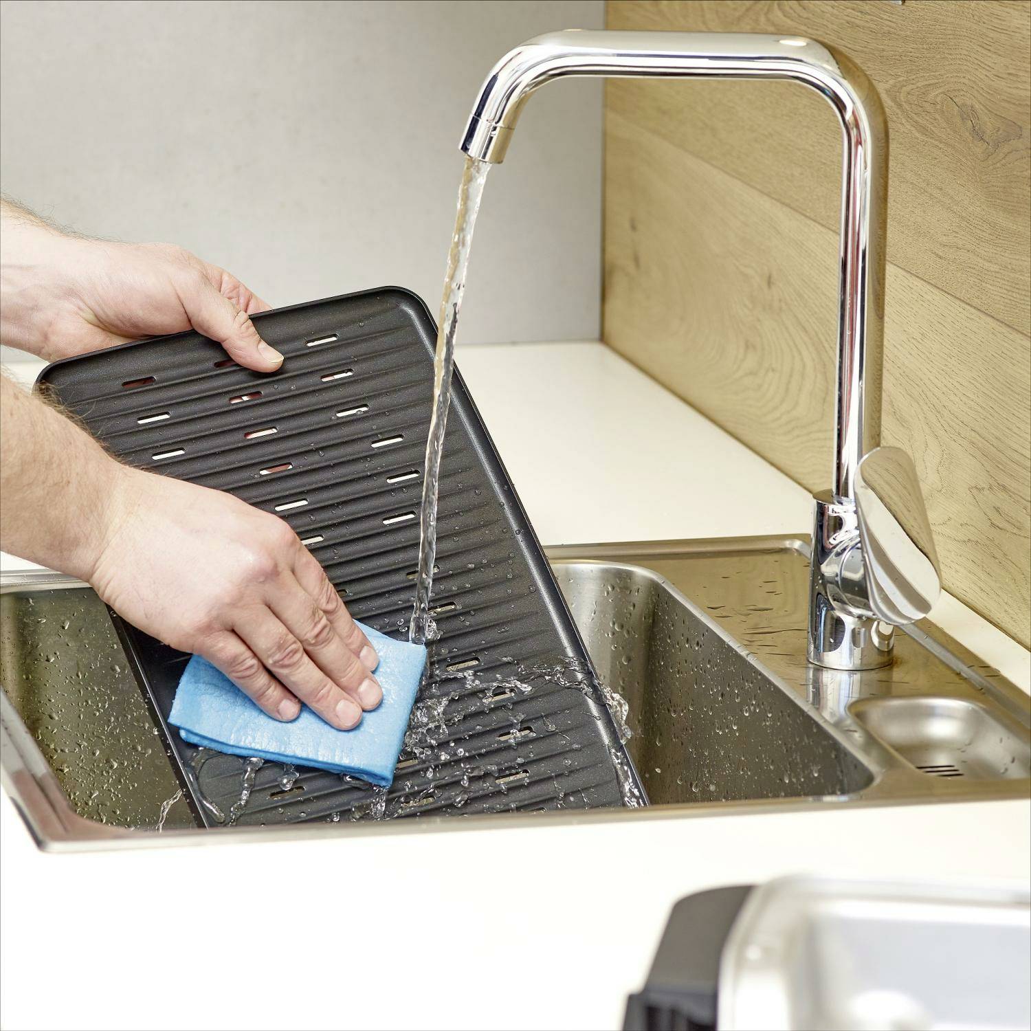 Une personne rince une égouttoir avec une éponge bleue sous l'eau courante dans un évier de cuisine moderne.