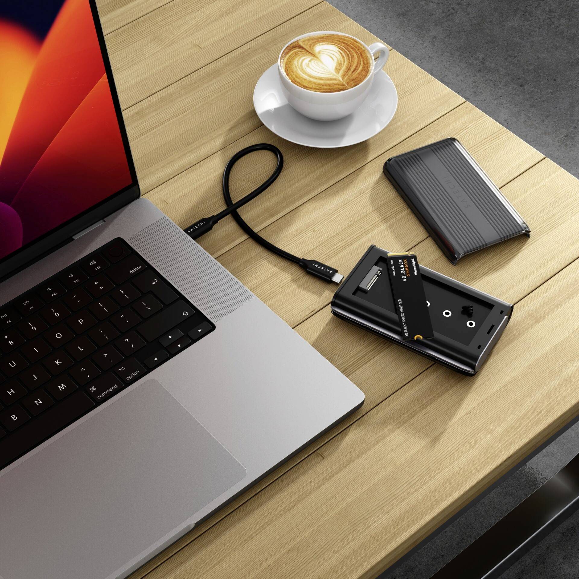 Sur une table en bois sont posés un ordinateur portable, une tasse de cappuccino, un disque dur externe, un lecteur de cartes et un câble USB.