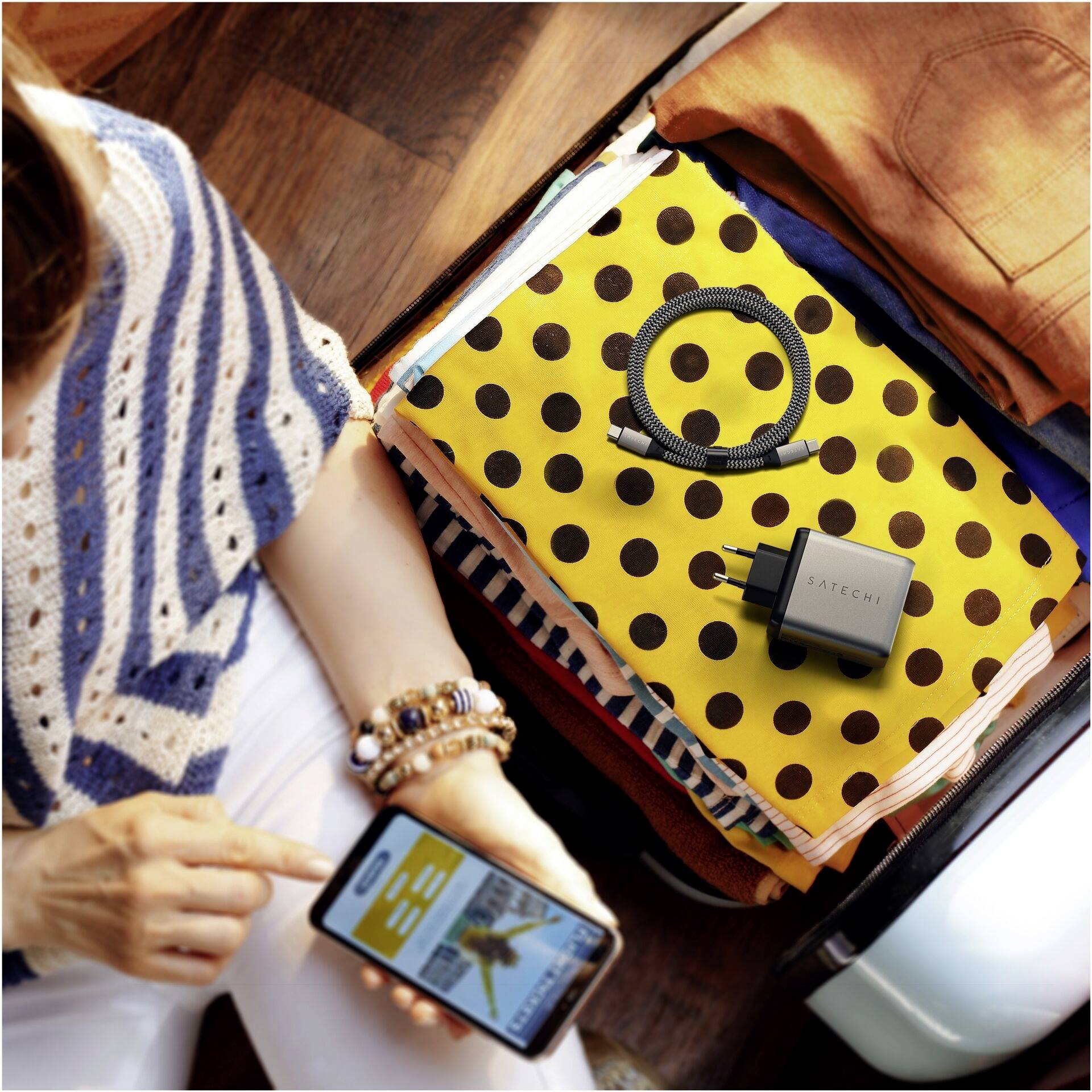Une personne prépare sa valise, regardant son smartphone avec une application, un tissu jaune à pois noirs dans la valise ; un câble de charge et un adaptateur à côté.