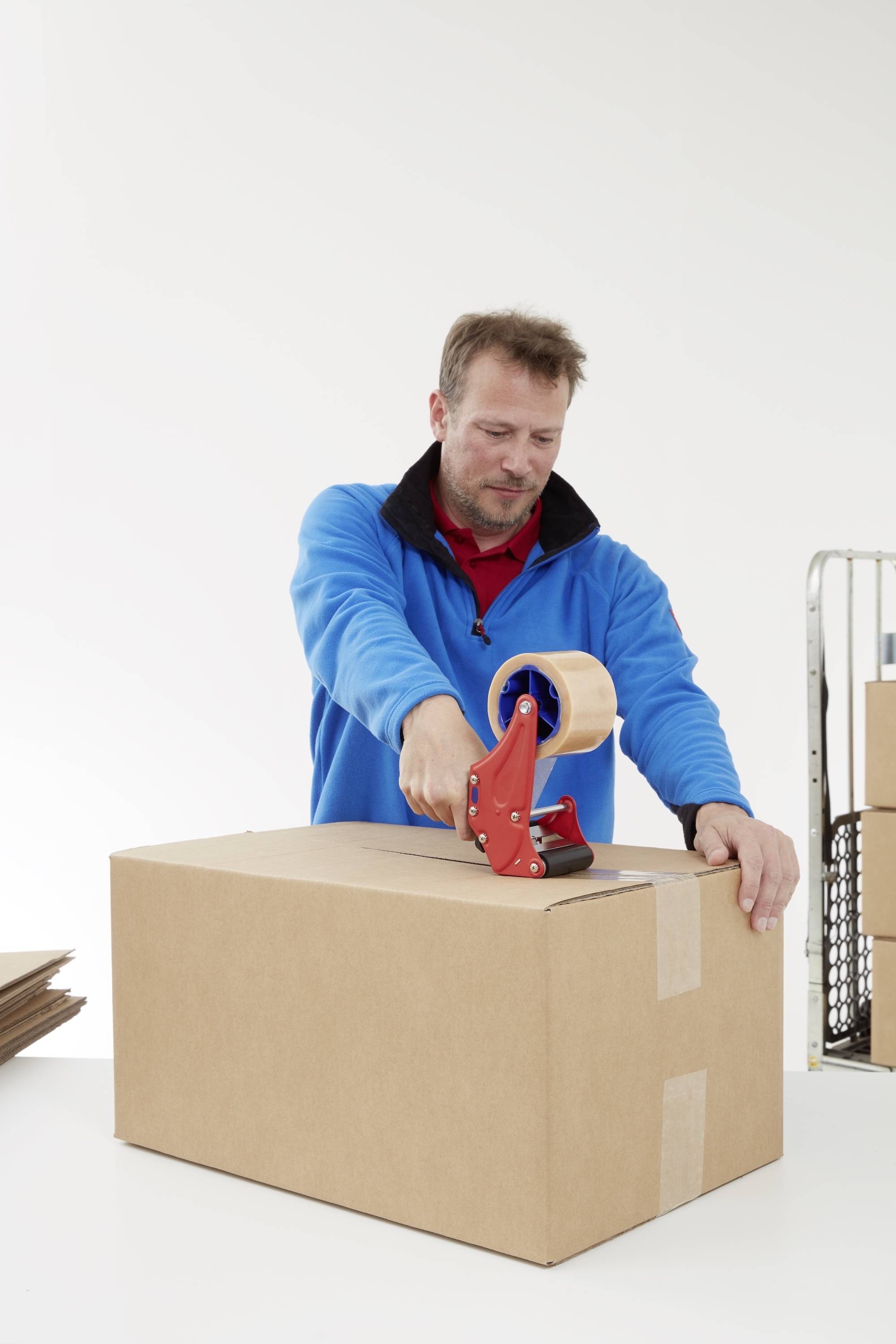 Un homme portant une veste bleue referme un carton avec du ruban adhésif. D'autres cartons sont visibles en arrière-plan.