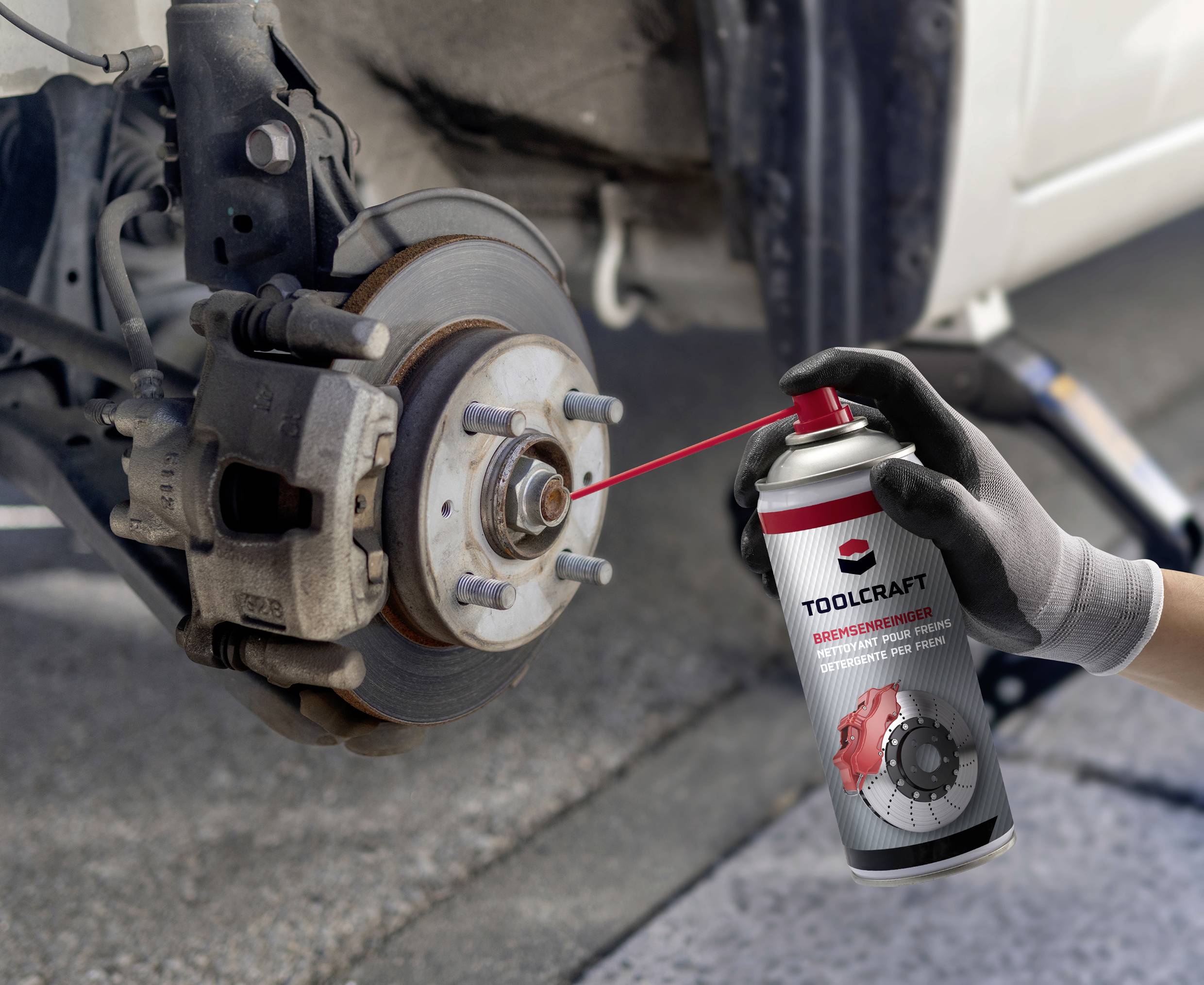 Une main vaporise un lubrifiant sur un frein de véhicule automobile pour effectuer une maintenance. Une voiture est surélevée sur un pont dans le fond.