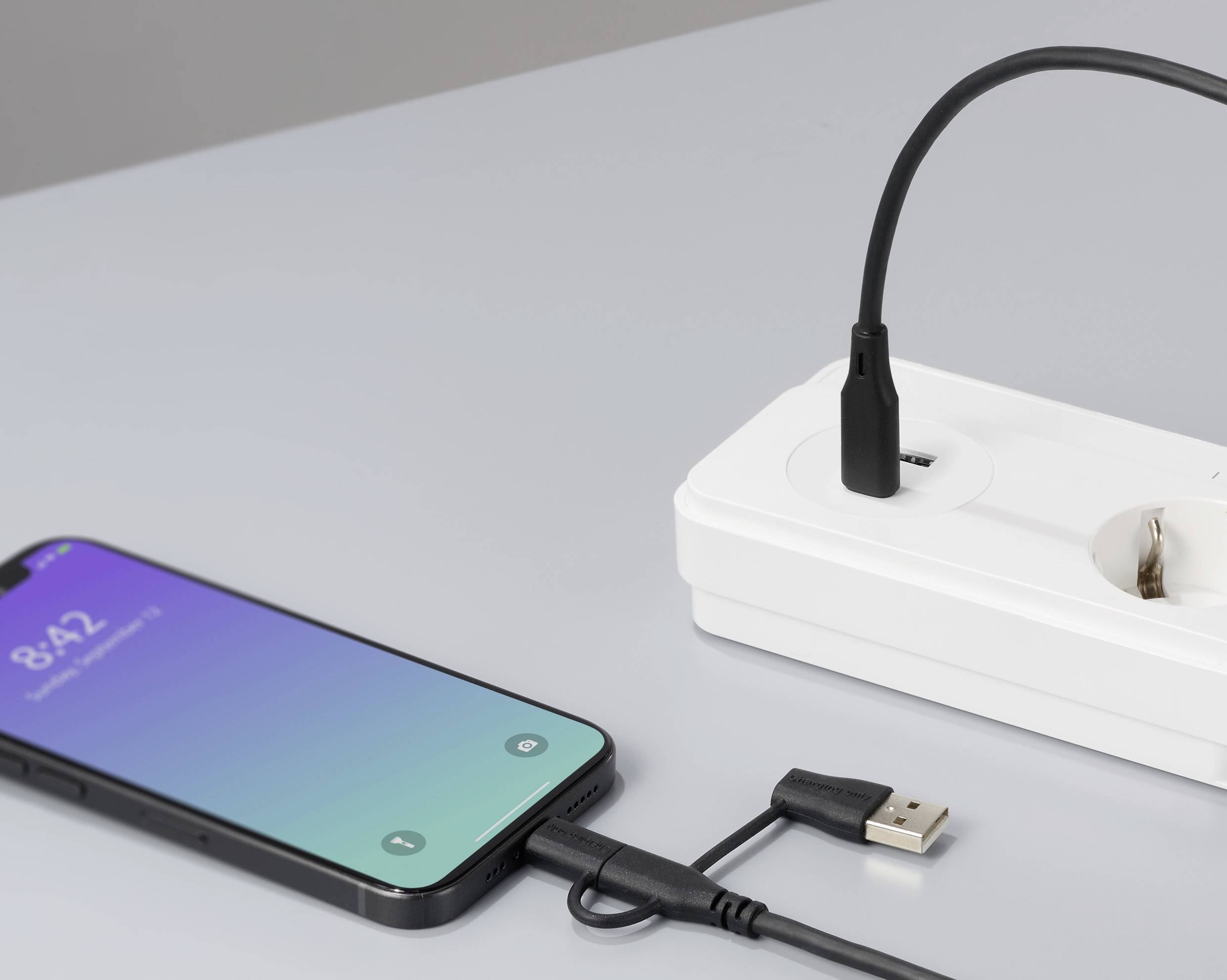 Un smartphone est en charge via un câble USB branché à une prise électrique. L'écran affiche l'heure 8:42.