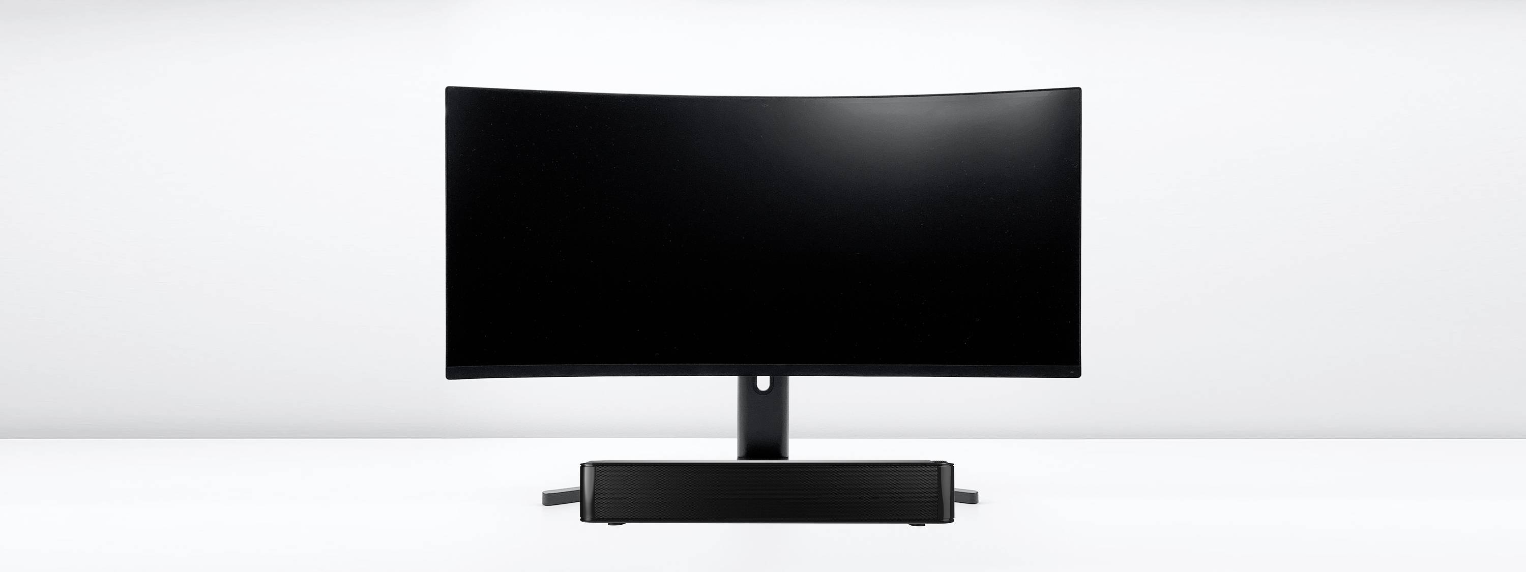 Un écran d'ordinateur incurvé noir posé sur une table blanche, avec une barre de son noire en dessous, sur un fond gris.