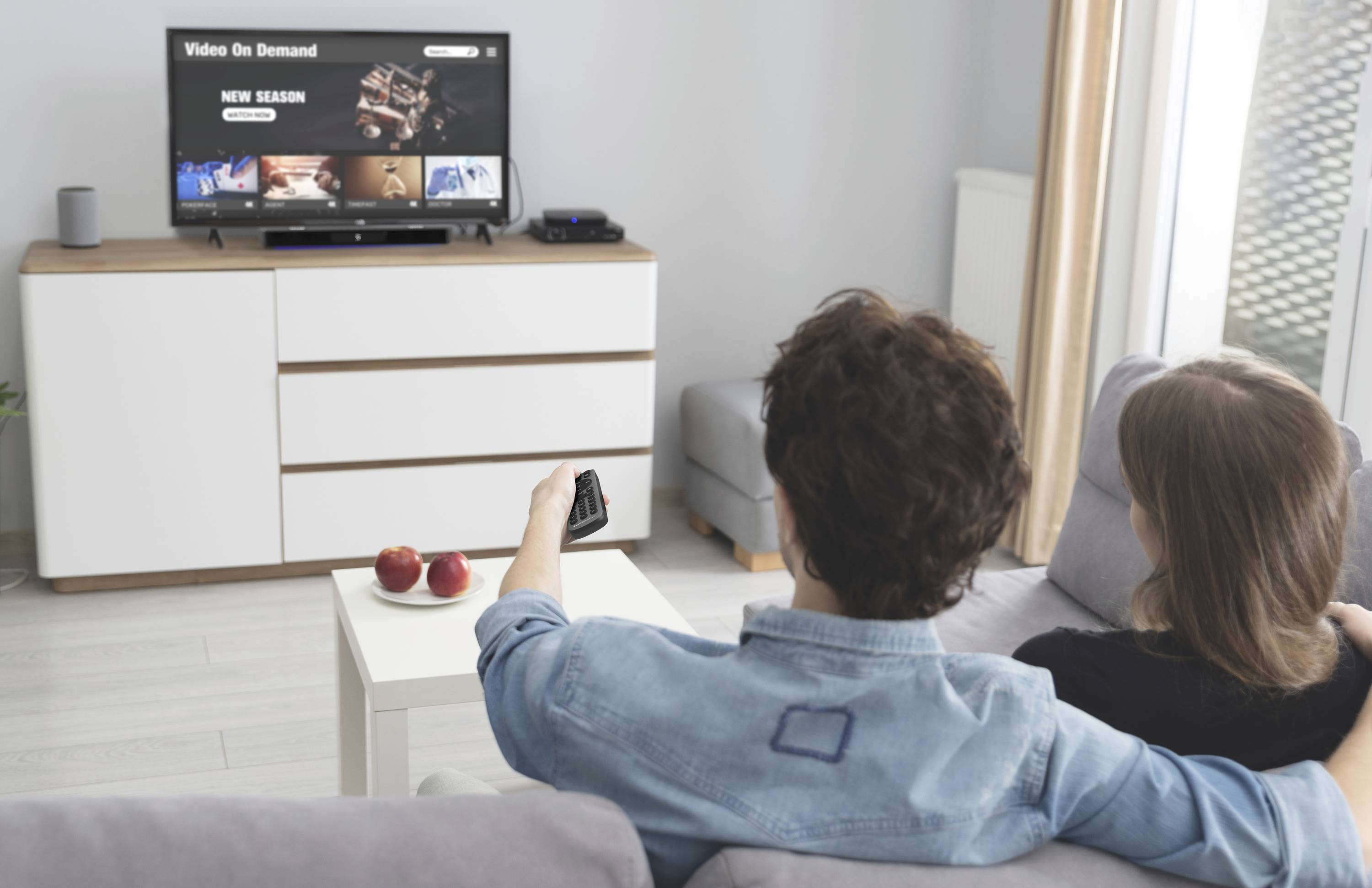 Un couple est assis sur un canapé et regarde la télévision. Sur l'écran, une option de vidéo à la demande est visible.