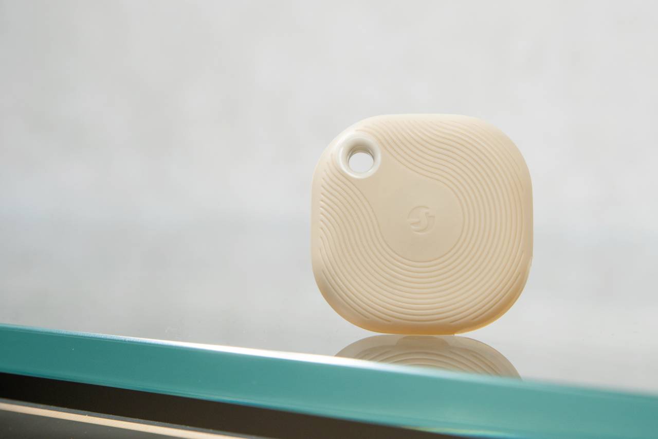 Un tracker beige, carré, aux coins arrondis, avec un trou pour cordon dans le coin supérieur, est posé sur une surface en verre.