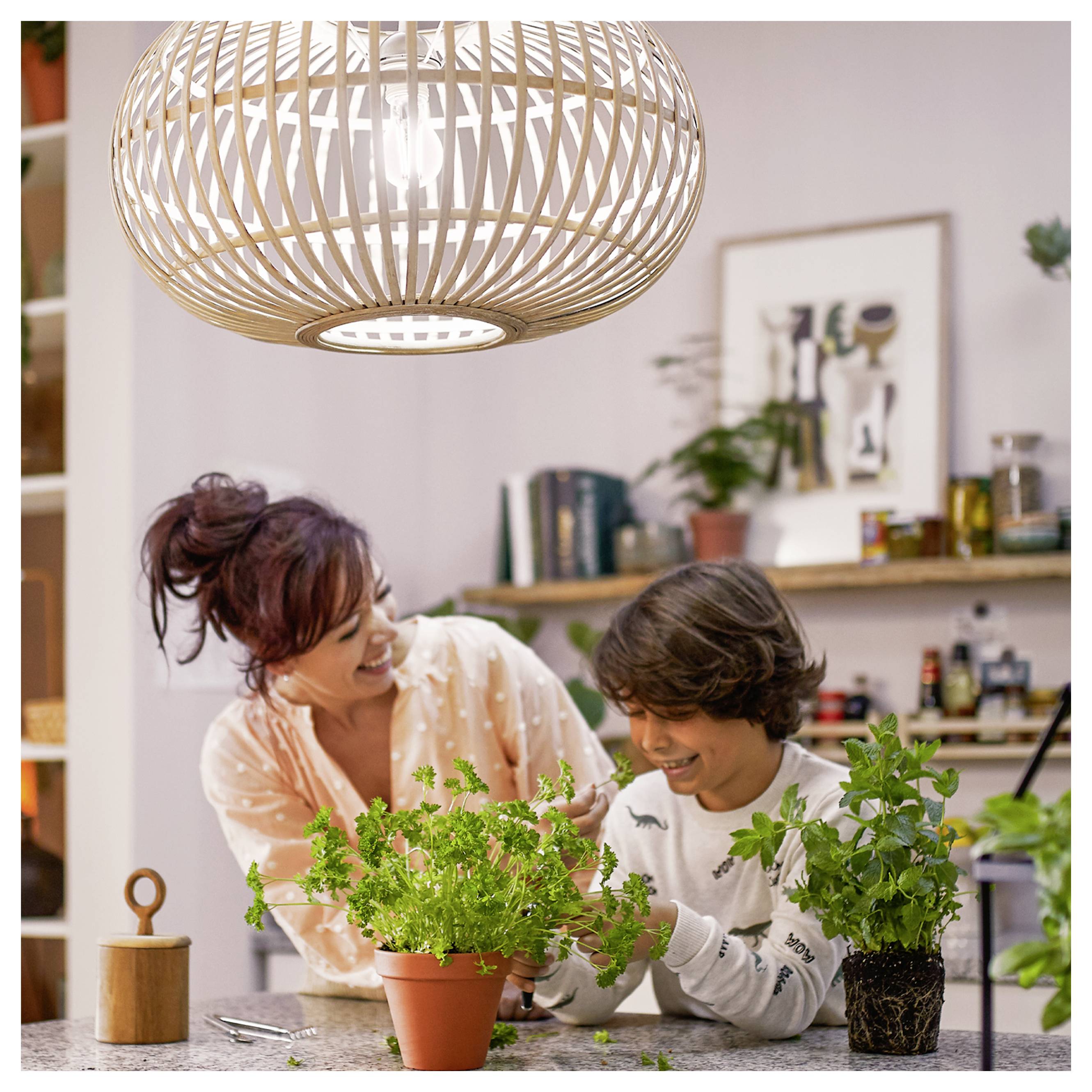 Femme et garçon s'occupant des herbes en pot sur un plan de travail de cuisine, souriants ensemble sous une lampe suspendue.