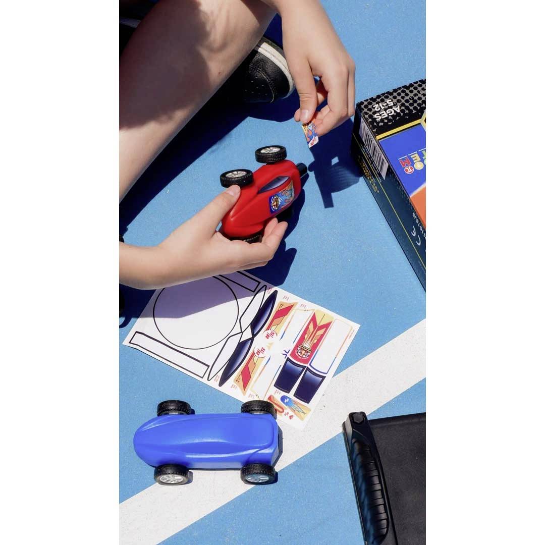 Un enfant sur un fond bleu colle des autocollants sur une voiture jouet rouge. Une voiture bleue et des feuilles d'autocollants sont posées à côté.
