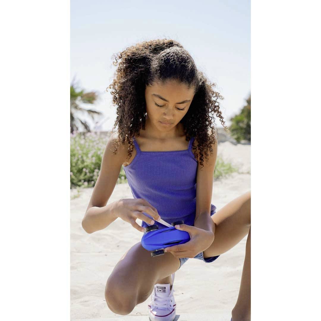 Une fille agenouillée sur la plage, prenant quelque chose dans un récipient bleu ; des palmiers et des arbres sont visibles en arrière-plan.