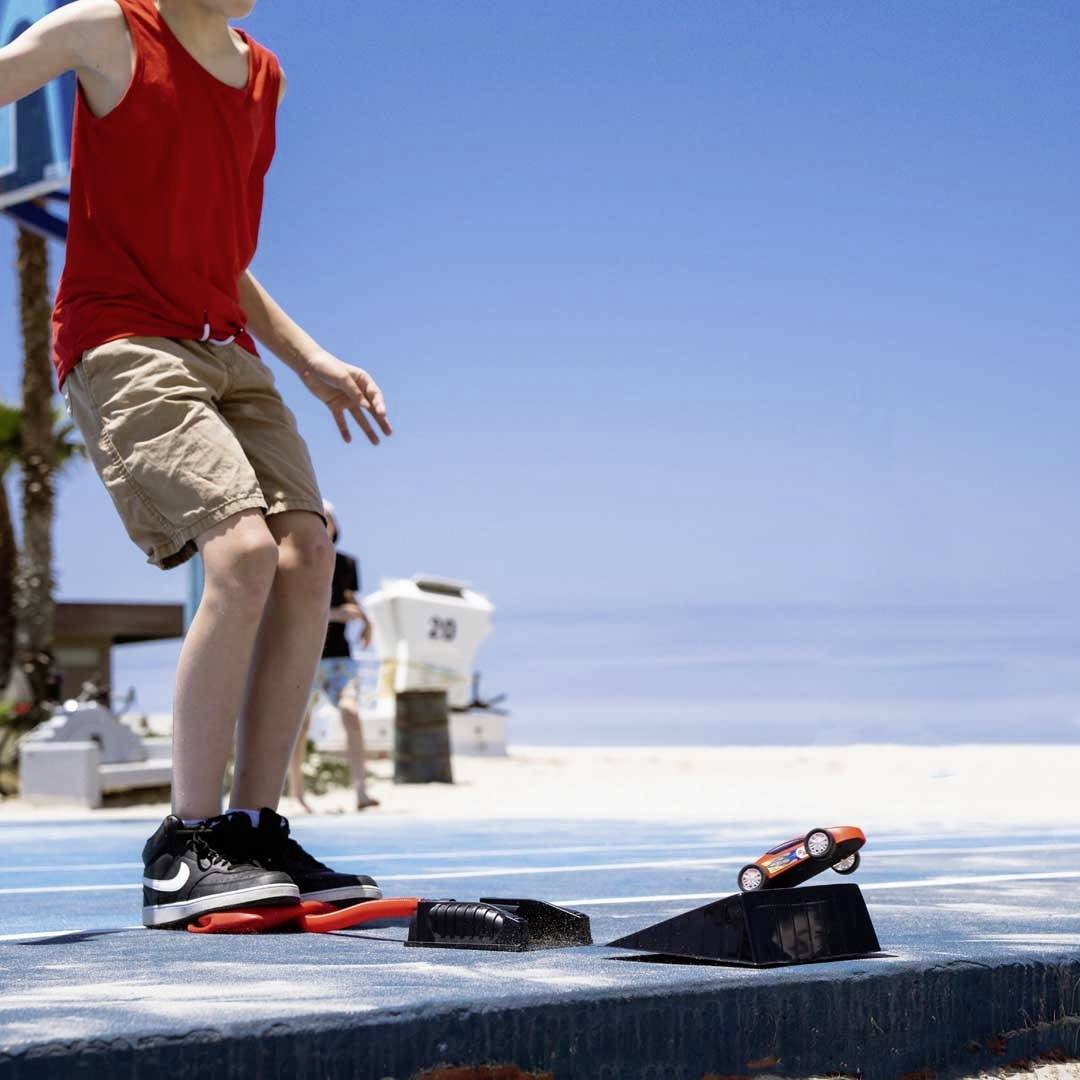 Un enfant portant un haut rouge et un short beige fait du skateboard sur une rampe près de la plage. Des palmiers sont visibles en arrière-plan.