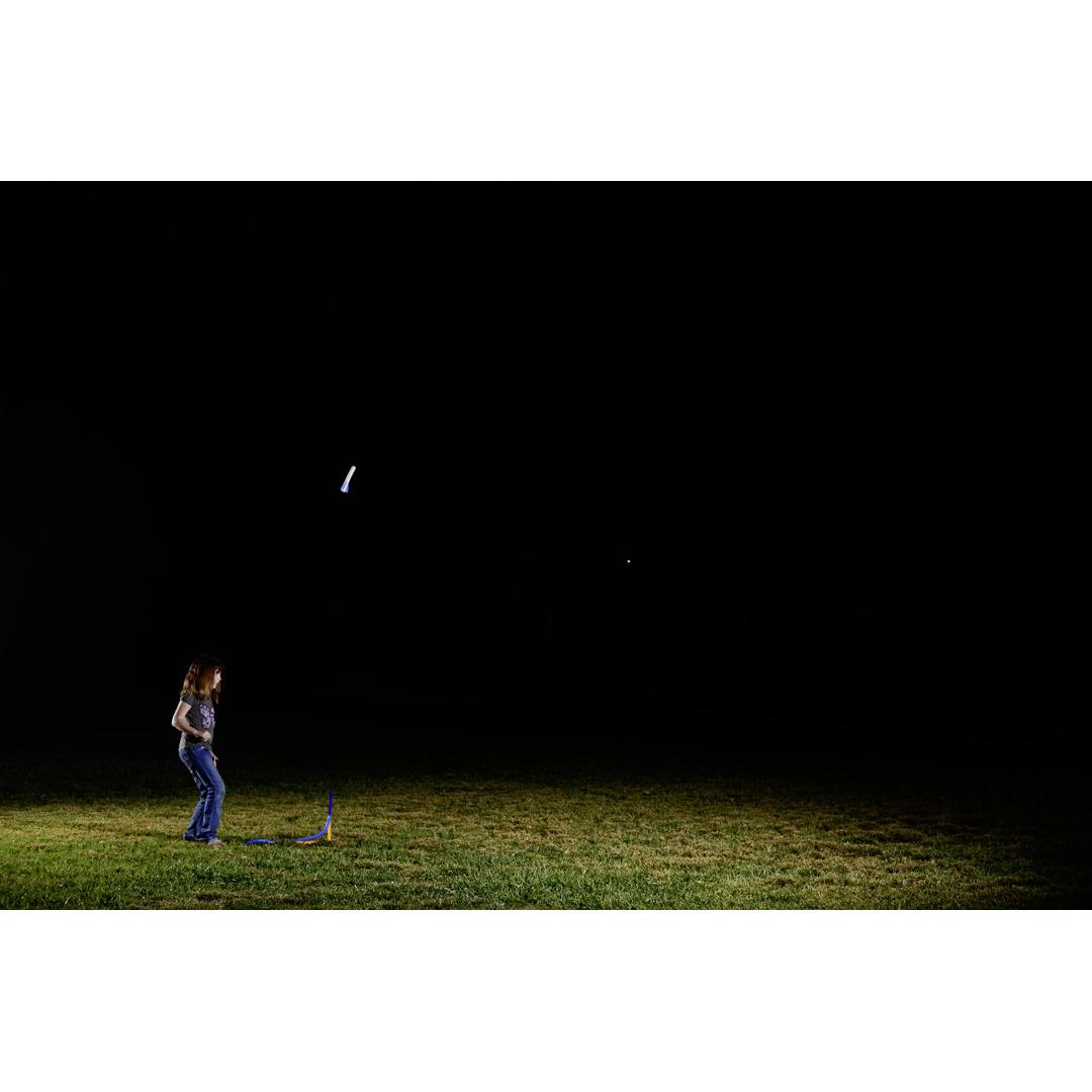 Une personne observe le lancement d'une fusée-modèle dans un champ pendant la nuit. La fusée illumine le ciel.